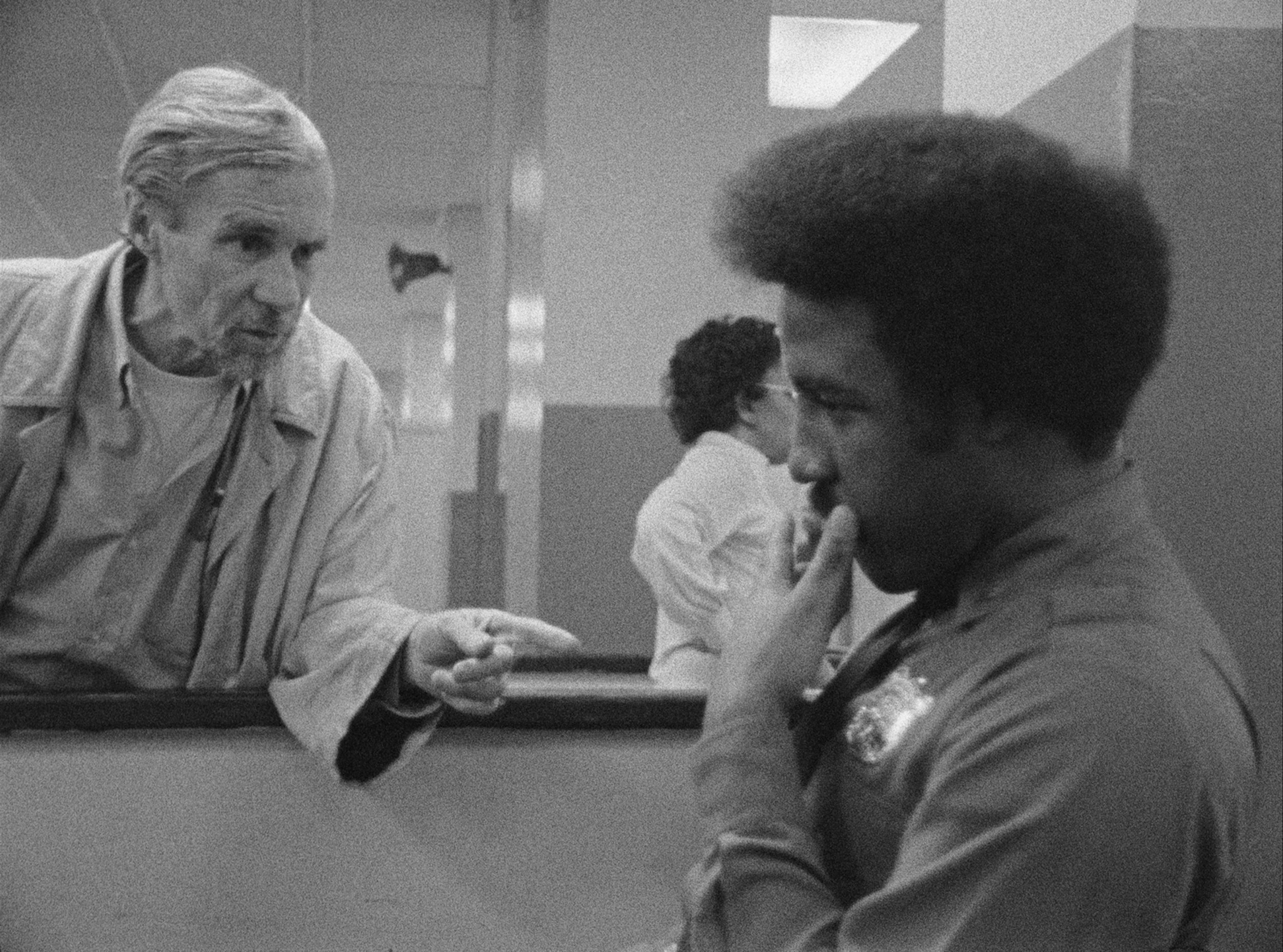 A black and white image of two men in a discussion, with one man pointing and the other listening intently. The background shows a person walking.