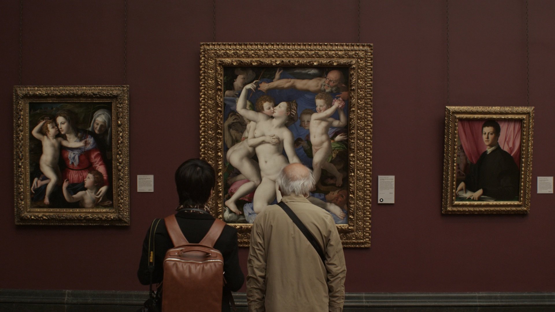 Two people viewing classic paintings in a museum, featuring portraits and mythological themes.
