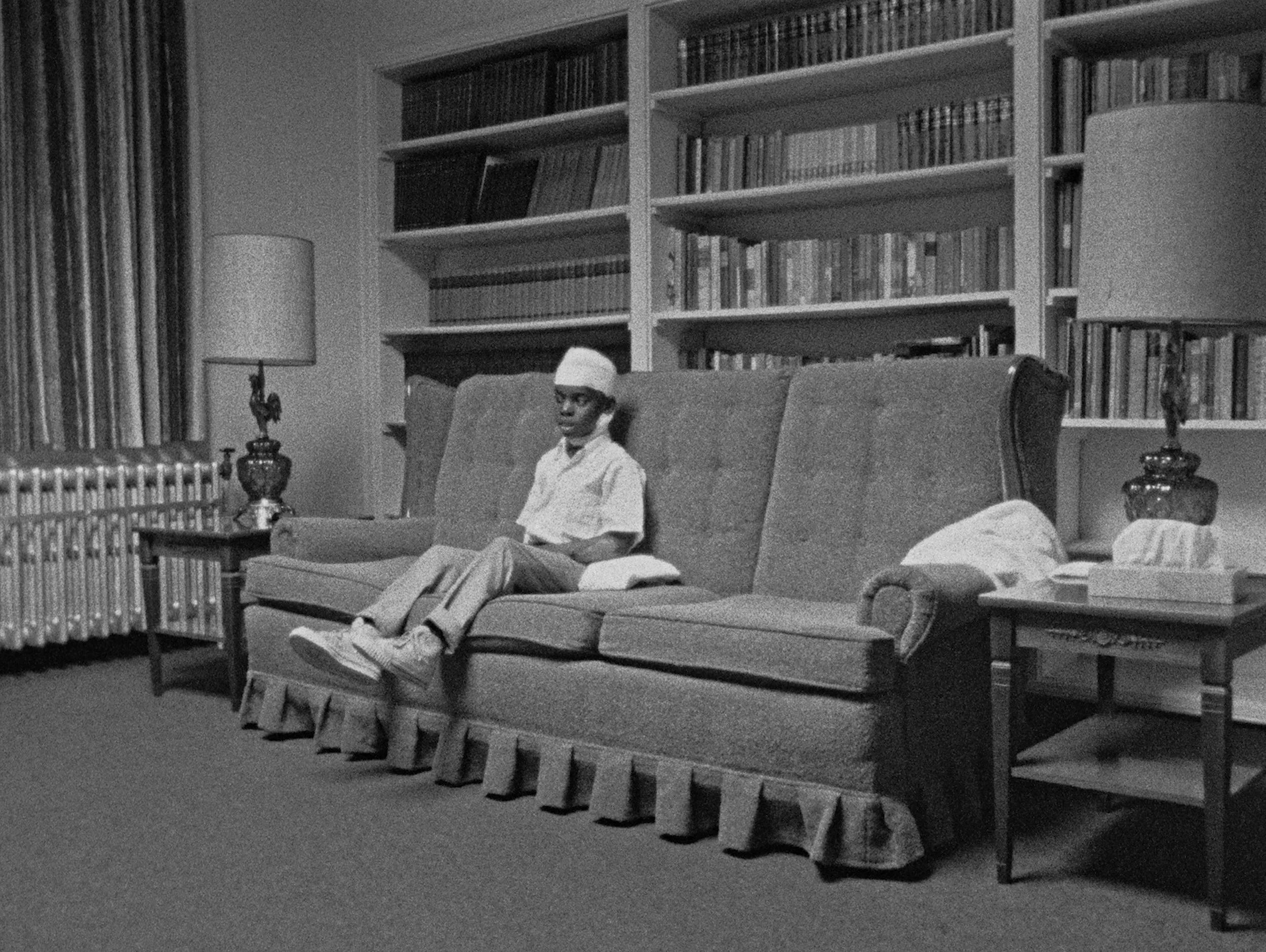 Black and white photo of a person sitting on a couch in a room with shelves full of books. The room has two lamps on side tables and a radiator on the left.