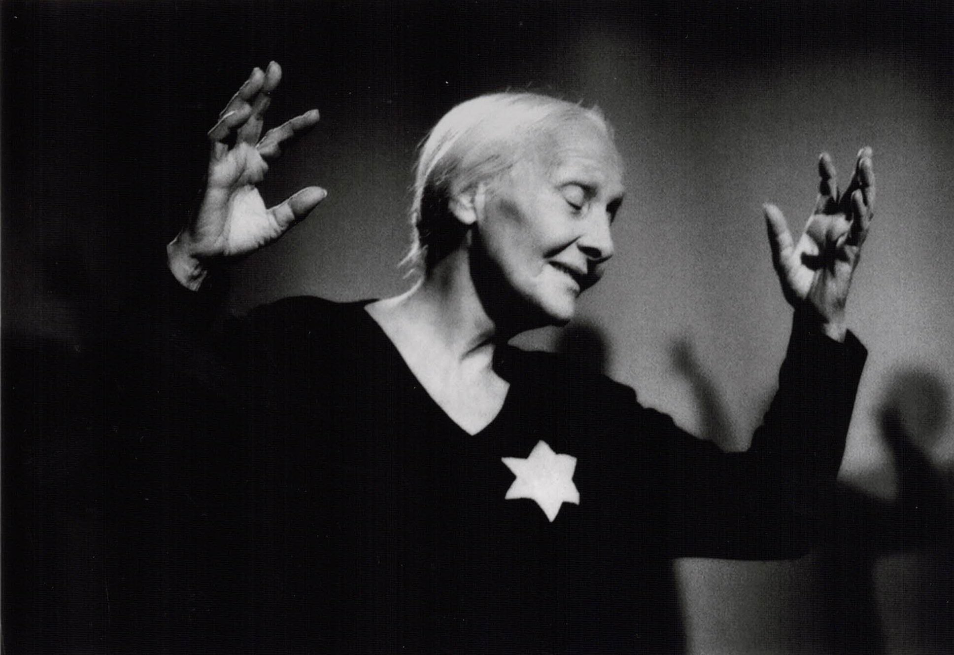 Elderly woman dancing, wearing dark clothes with a star-shaped emblem, in a black-and-white photo.