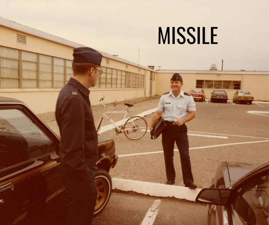 Two men in military uniforms conversing in a parking lot with cars and a bicycle, captioned "MISSILE".