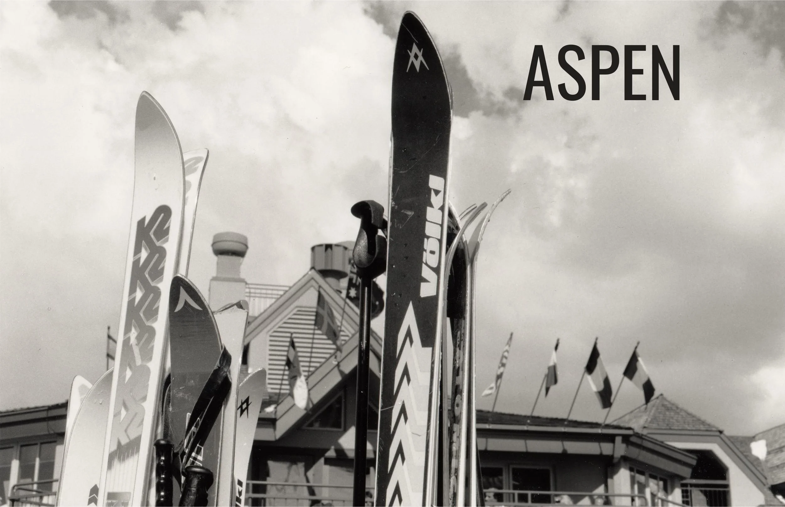 Black and white photo of ski poles and skis in Aspen with a building and flags in the background.