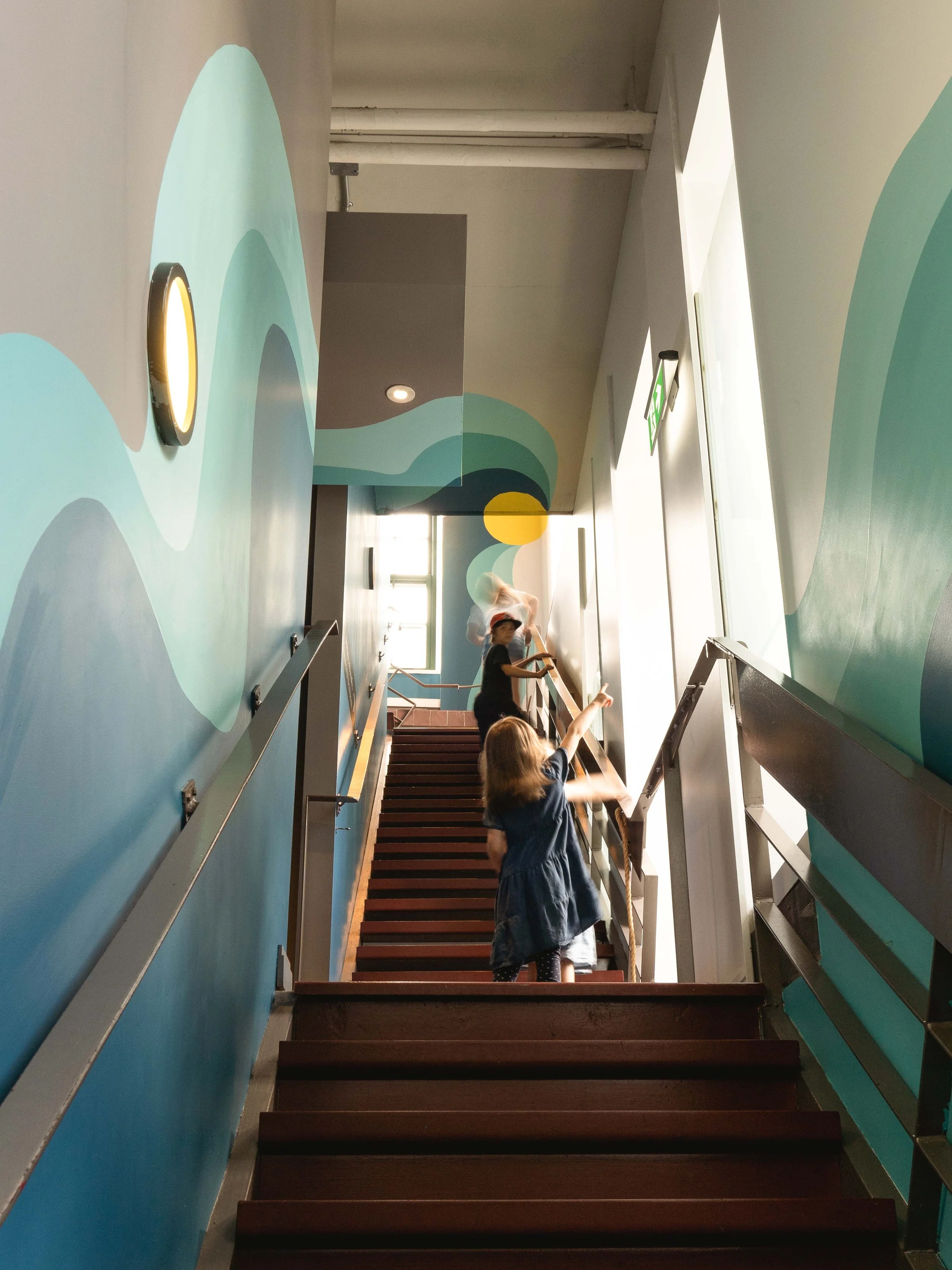 Design de la murale de l'escalier menant à la salle éducative. Cette murale parle du mouvement du fleuve et de l'histoire de Montréal. Cette murale a été co-créé par MU, La camaraderie et inscape