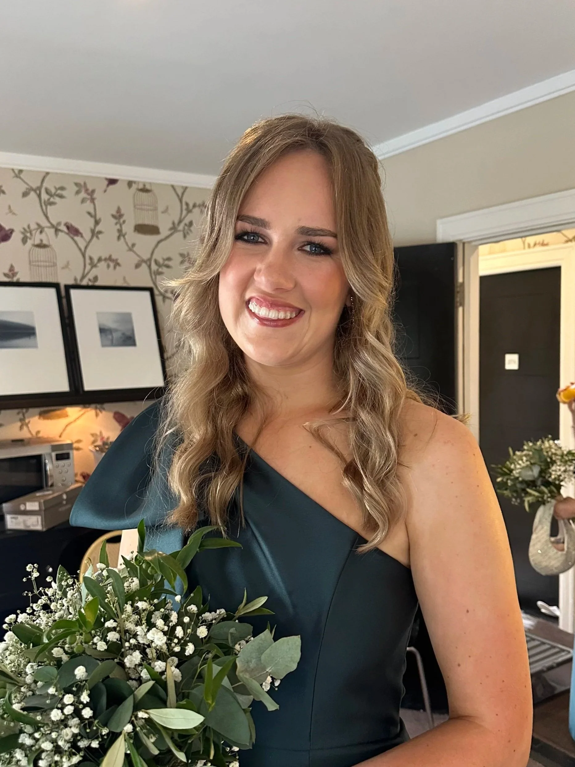 A woman in a dark teal dress holding a bouquet of white flowers, smiling in a room decorated for a celebration.