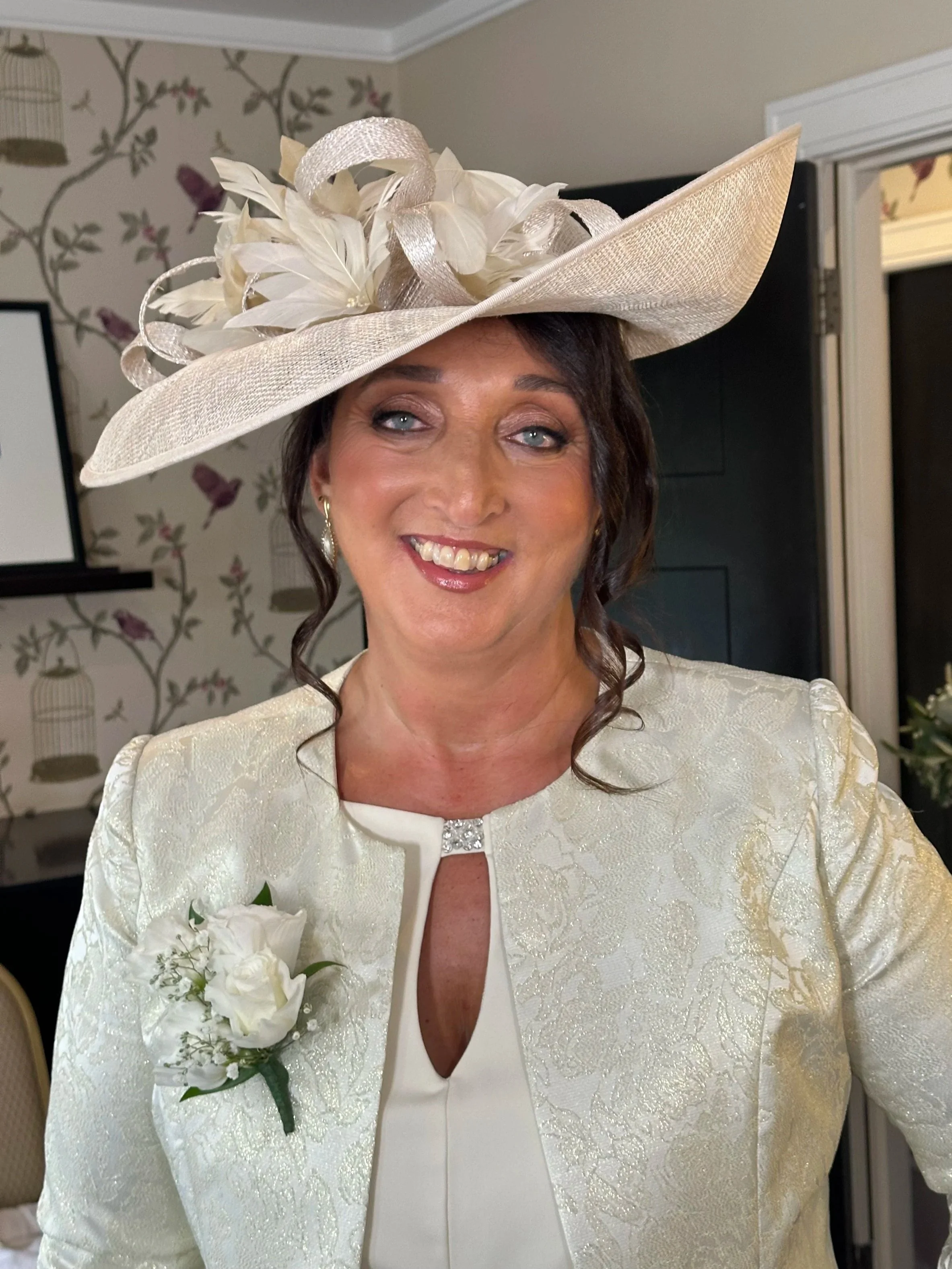 A woman wearing a large, cream-colored fancy hat with feathers and ribbons, a cream jacket with floral pattern, and a white top with a keyhole neckline, smiling indoors.