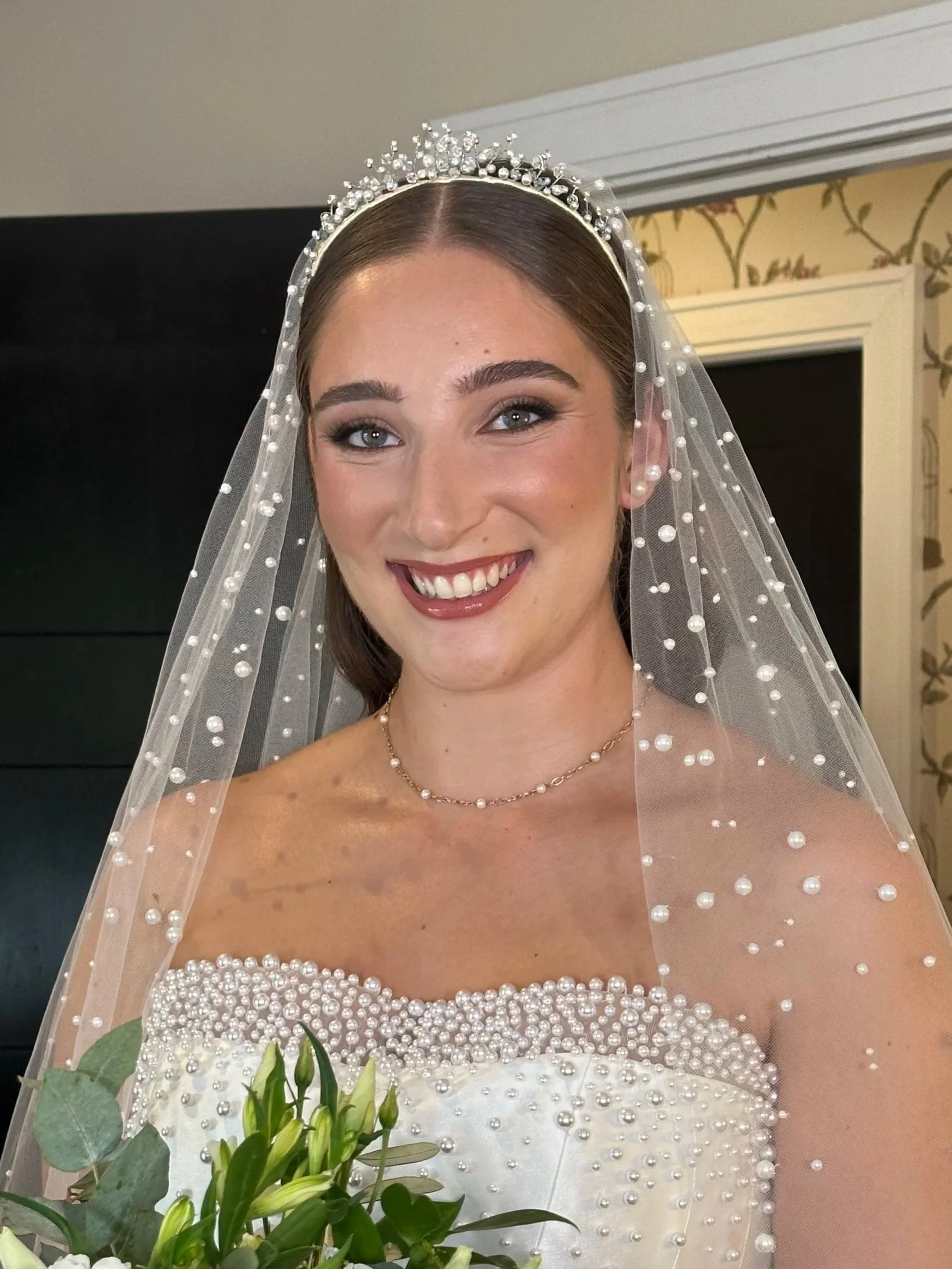 A smiling bride holding a bouquet of white flowers, wearing a pearl-embellished wedding dress and veil, with a tiara and jewelry, standing indoors.