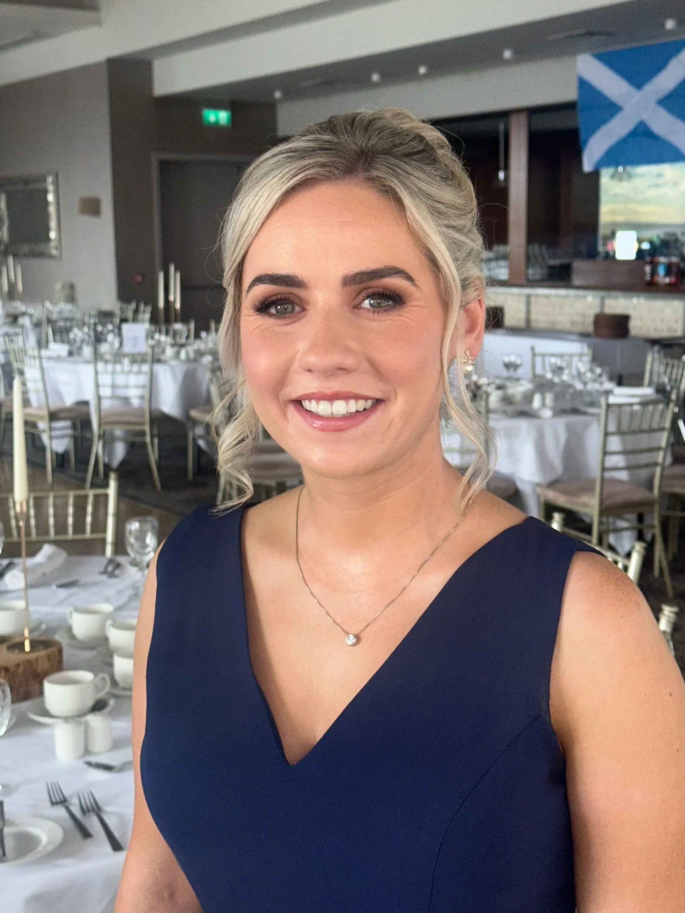 A woman in a blue dress is standing in a decorated event hall with tables set for a meal, and a flag with a blue and white cross is hanging in the background.