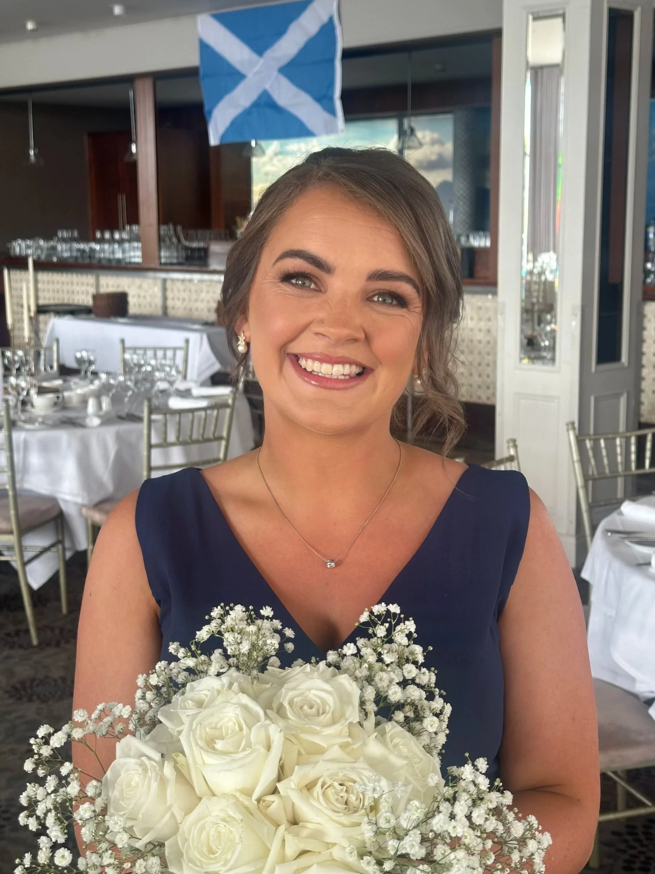 A young woman smiling and holding a bouquet of white roses, wearing a navy blue dress, in a decorated event space with tables and chairs, and a Scottish flag hanging in the background.
