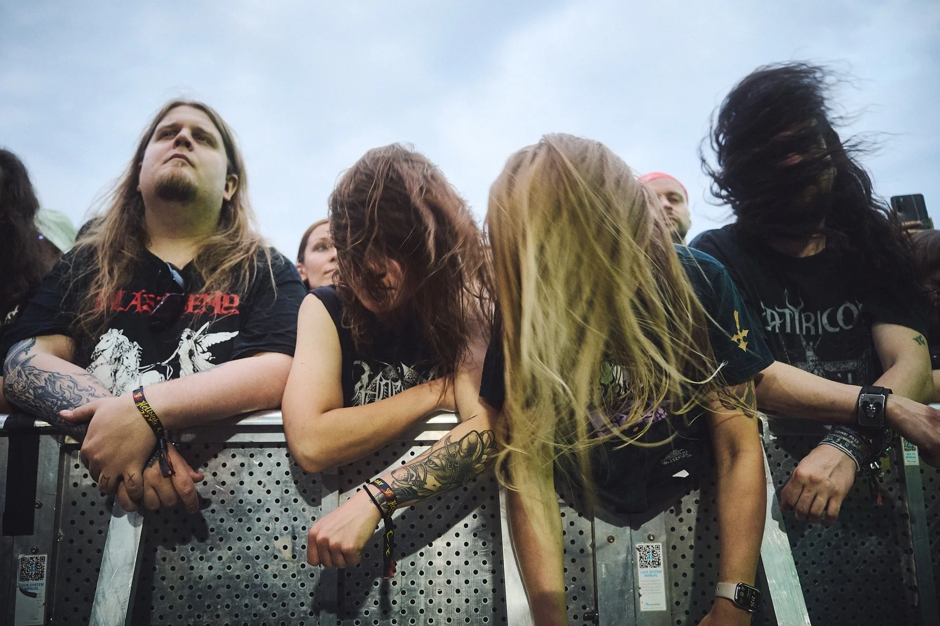 Ihmisiä rockkonsertissa nojaamassa metallikaiteeseen, päitä heiluttaen, pukeutuneina tummiin T-paitoihin.