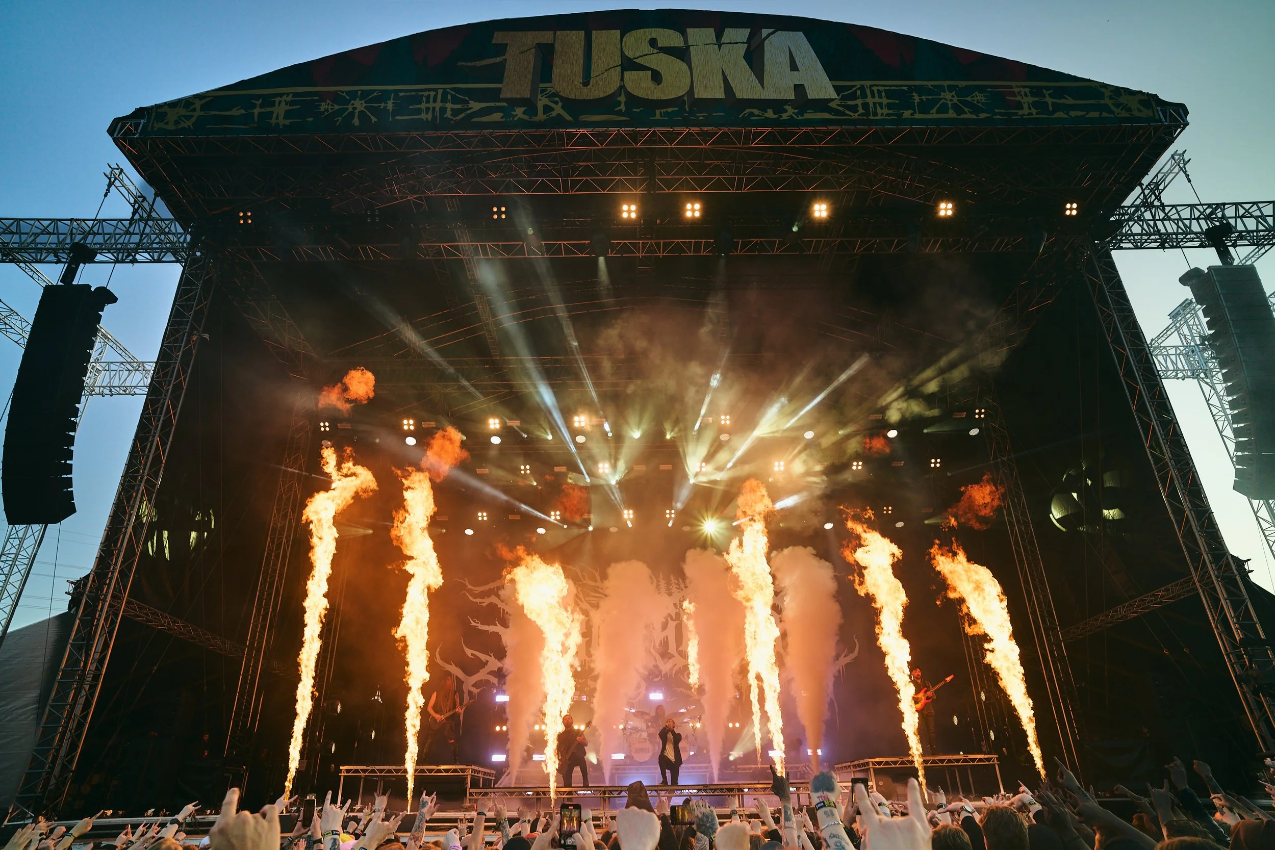 Suuri ulkoilmakonsertti lavalla, jossa esiintyjä ja bändi, taustalla liekin- ja valoefektejä ja katsomo täynnä ihmisiä, joista osa ottaa valokuvia ja videota.