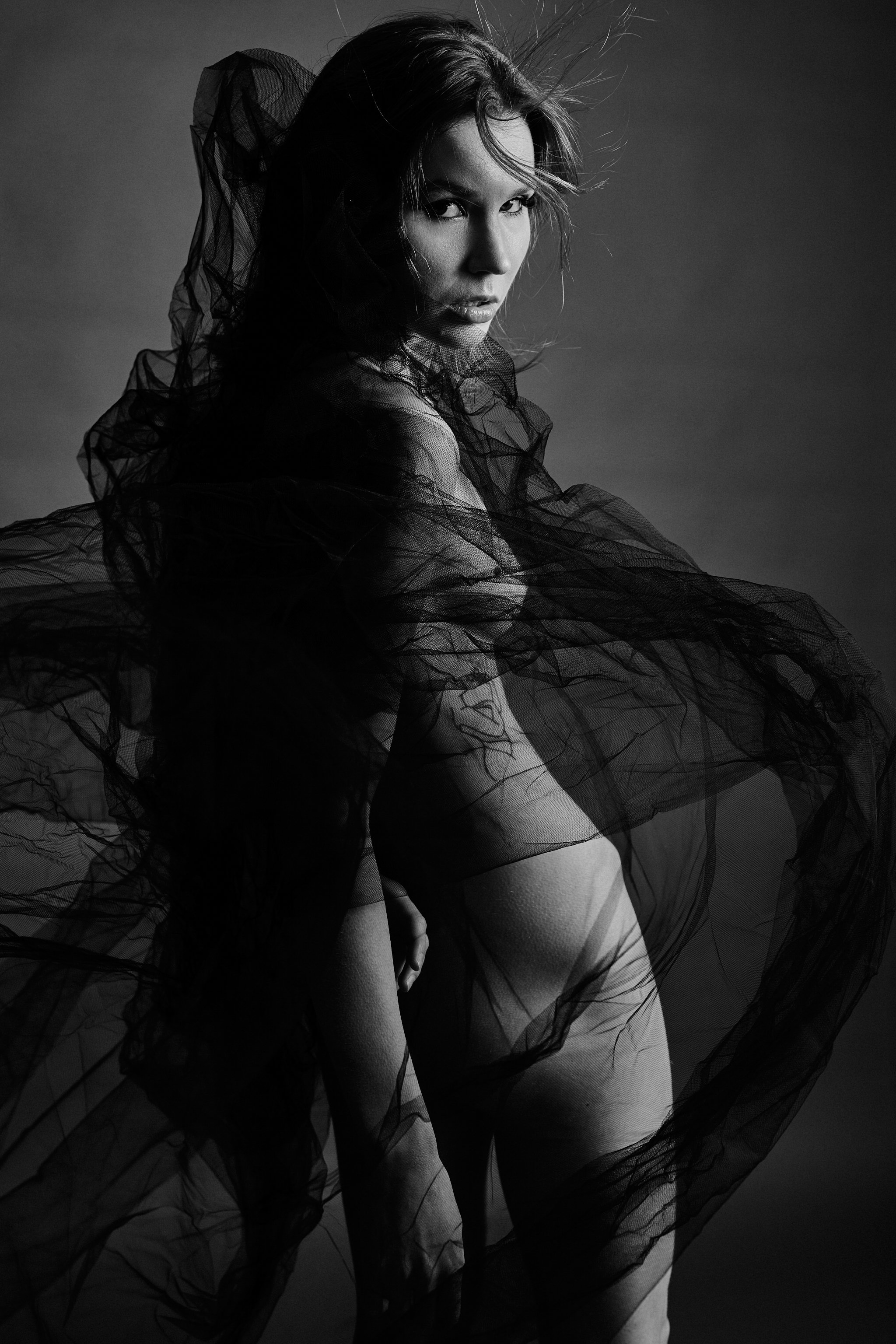 Musta-valkoinen valokuva naisesta, joka katsoo vakavana kameraan, osittain peitettynä mustasta, läpikuultavasta kankaasta.