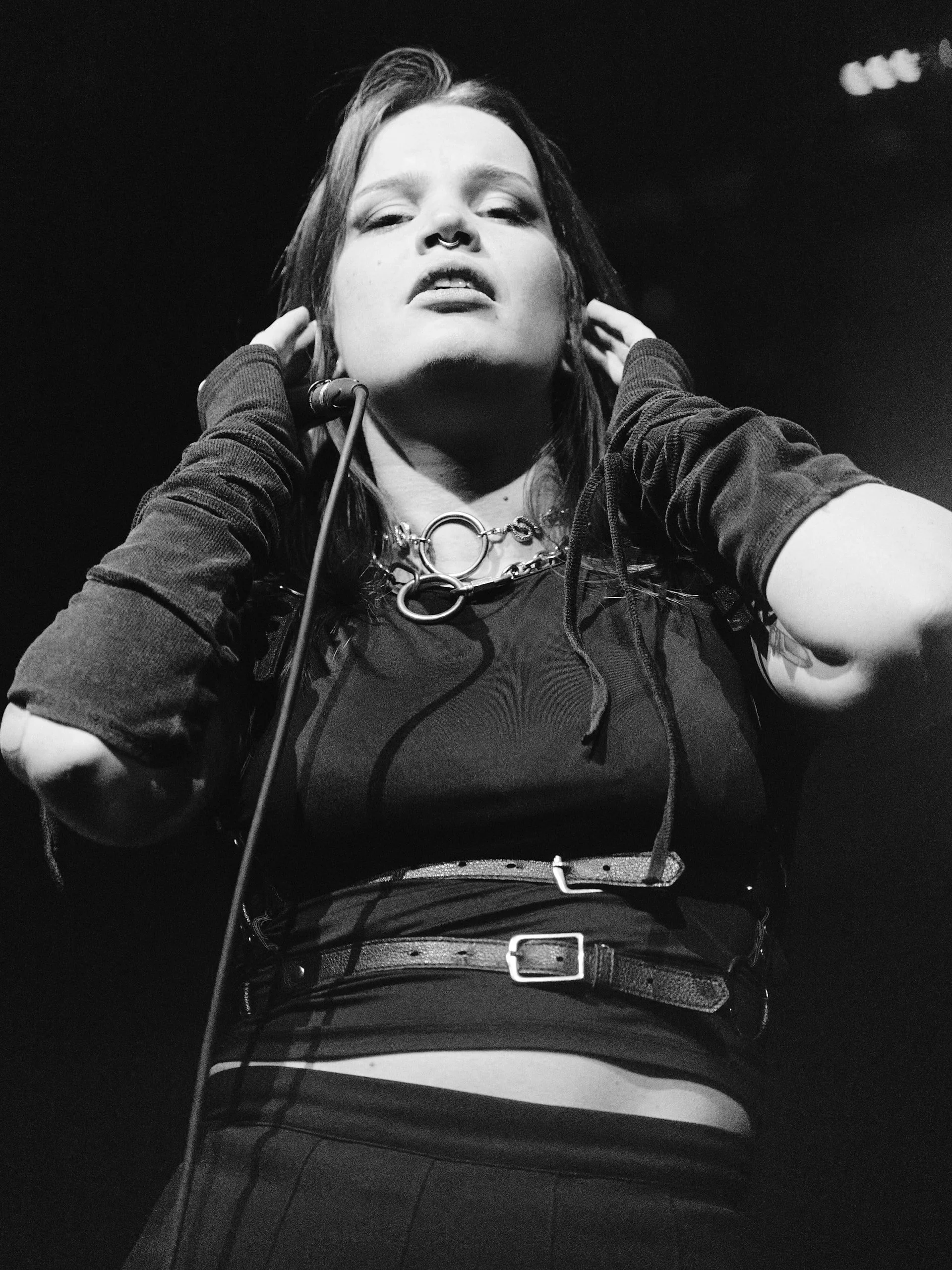 Henkilö esiintyy lavalla mikrofoni kädessään, pukeutunut tummiin vaatteisiin ja koruihin.