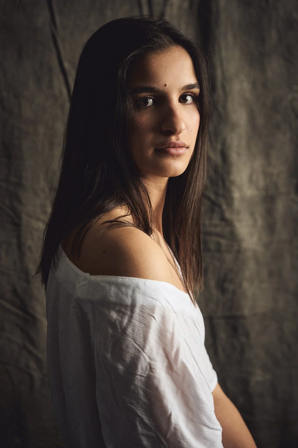 Valkoinen oleskelupaita, tummat hiukset, luonnollinen ilme, taustalla harmaa kiviseinä.