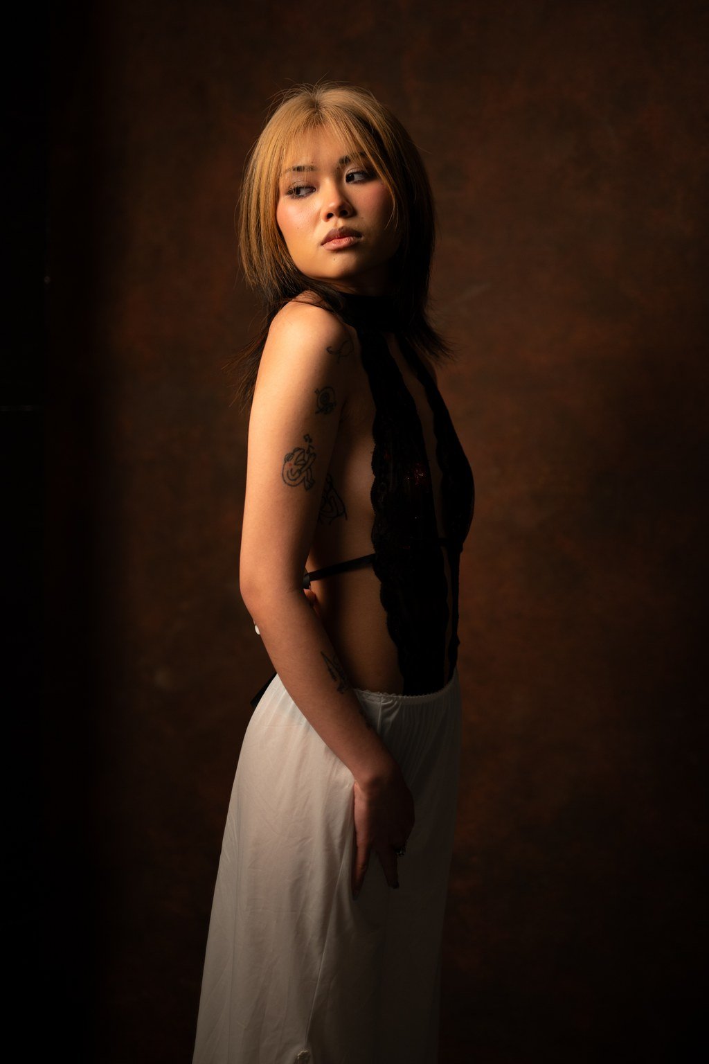 Nuori nainen seisoo pimeässä taustalla, katsellen pois oikealle, vartalo hieman kääntyneenä, yllään musta pusero ja valkoiset housut, tatuointeja vasemmassa käsivarressa.