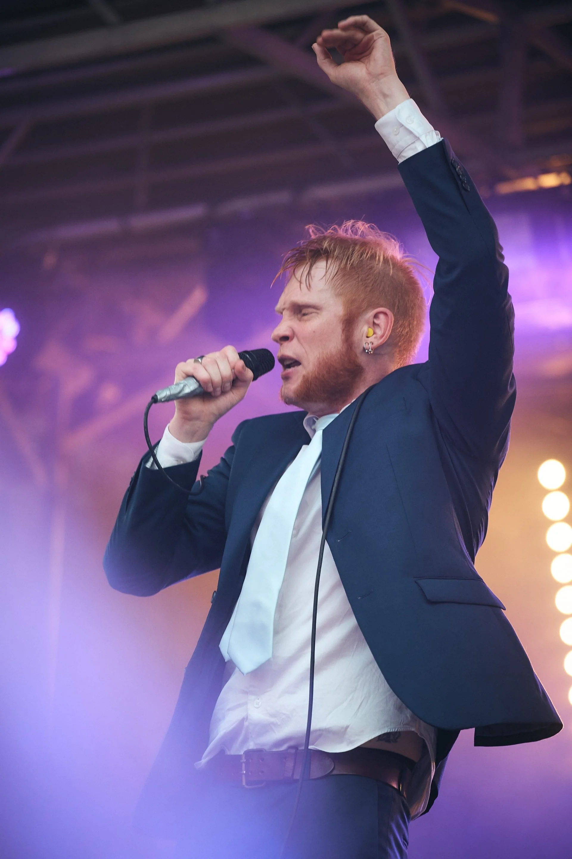 Mies laulaa mikrofonin kanssa konserttilavalla, pukeutuneena pukuun ja solmioon, toinen käsi ylhäällä.