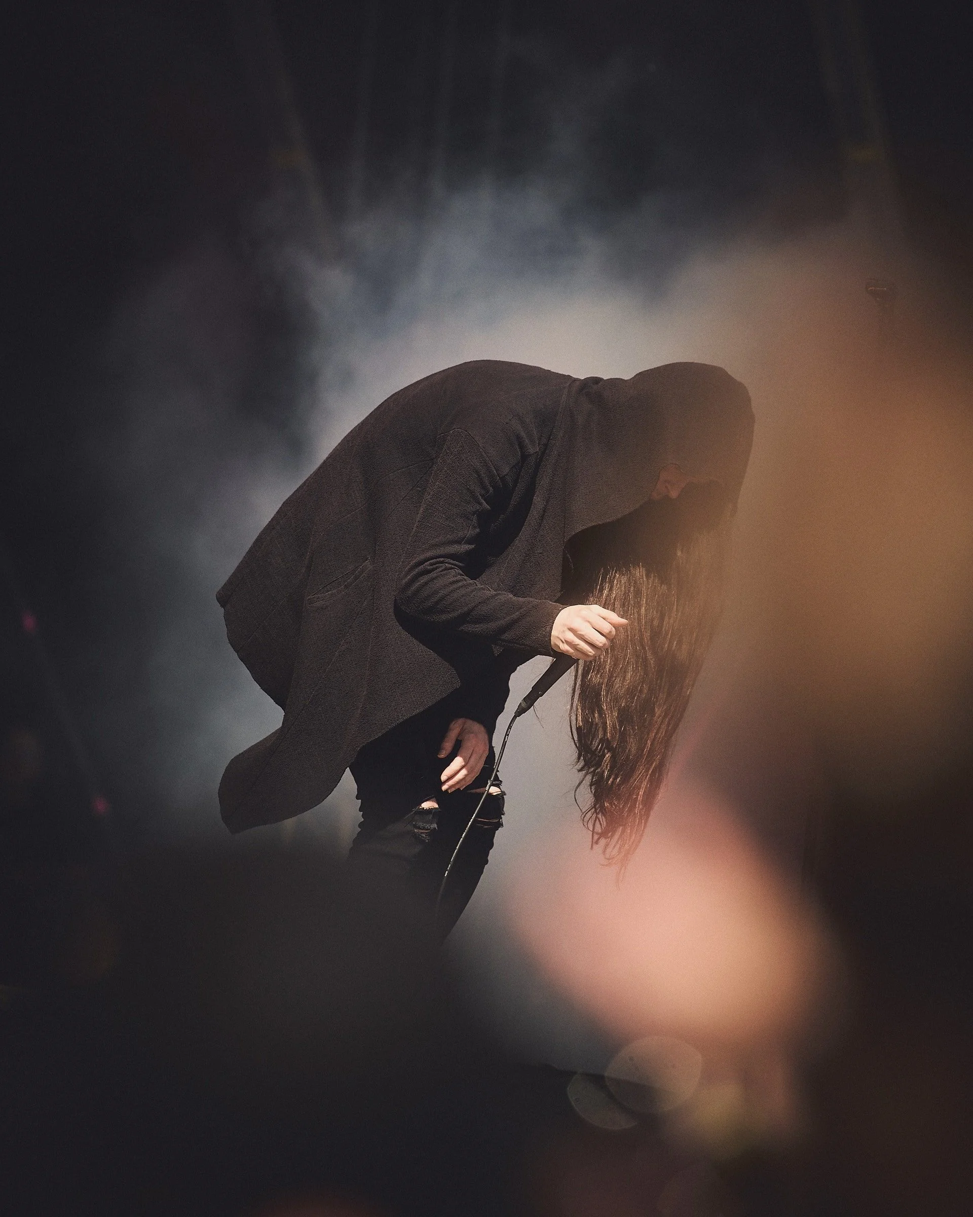 Henkilö pukeutuneena mustaan puseroon, kumartuu eteenpäin pitkät hiukset roikkuen. Kuvassa on sumuinen tausta ja keskittymistä valoon.