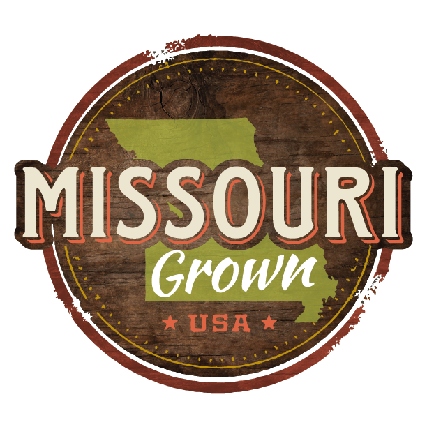 A circular wooden sign with text and graphics. It features the outline of the state of Missouri in green, the words "Missouri Grown" with "Missouri" in large block letters and "Grown" in script, and "USA" at the bottom with two stars, all on a rustic wood background.
