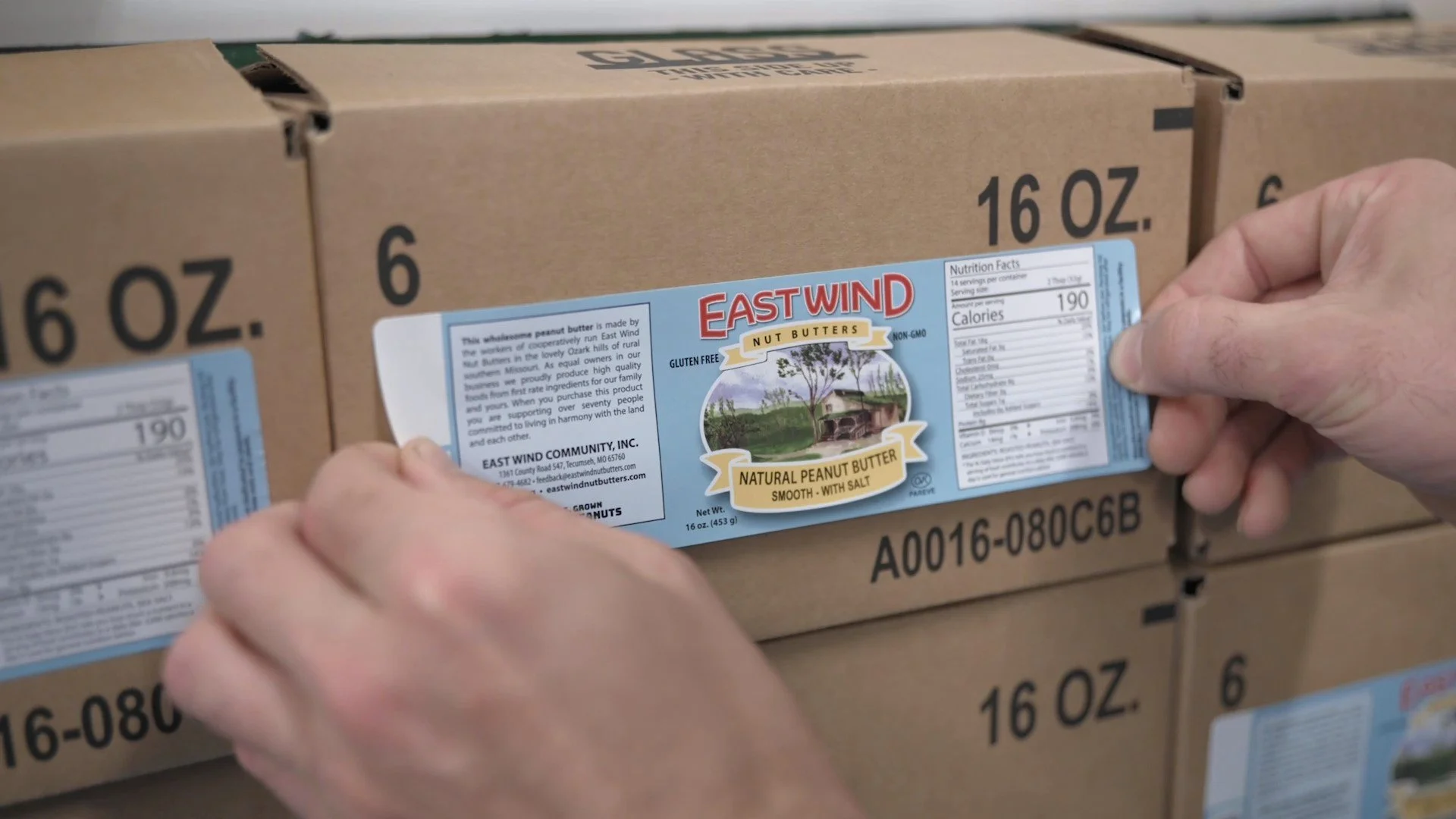 Close-up of a worker carefully applying the label to a shipping box containing jars of East Wind Smooth Valencia Peanut Butter with Salt
