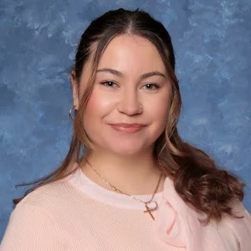 A young woman with brown hair styled in loose waves, wearing a light pink top and a gold necklace, smiling against a blue textured background.