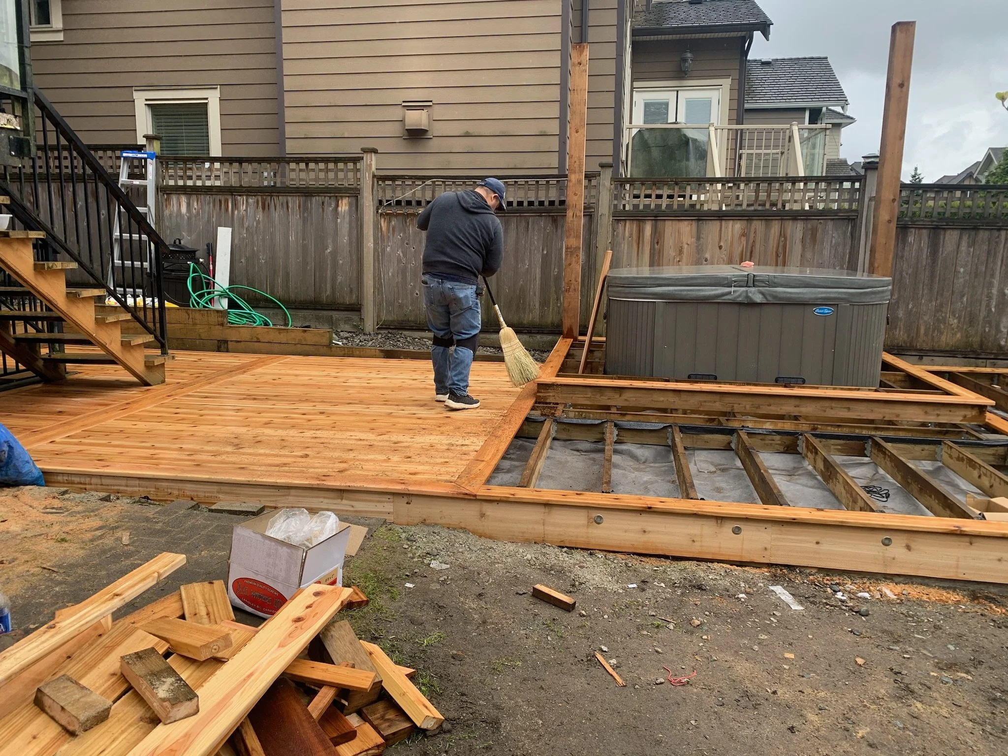 A person cleaning a freshly built wooden deck in a backyard, with a hot tub and a wooden fence in the background.