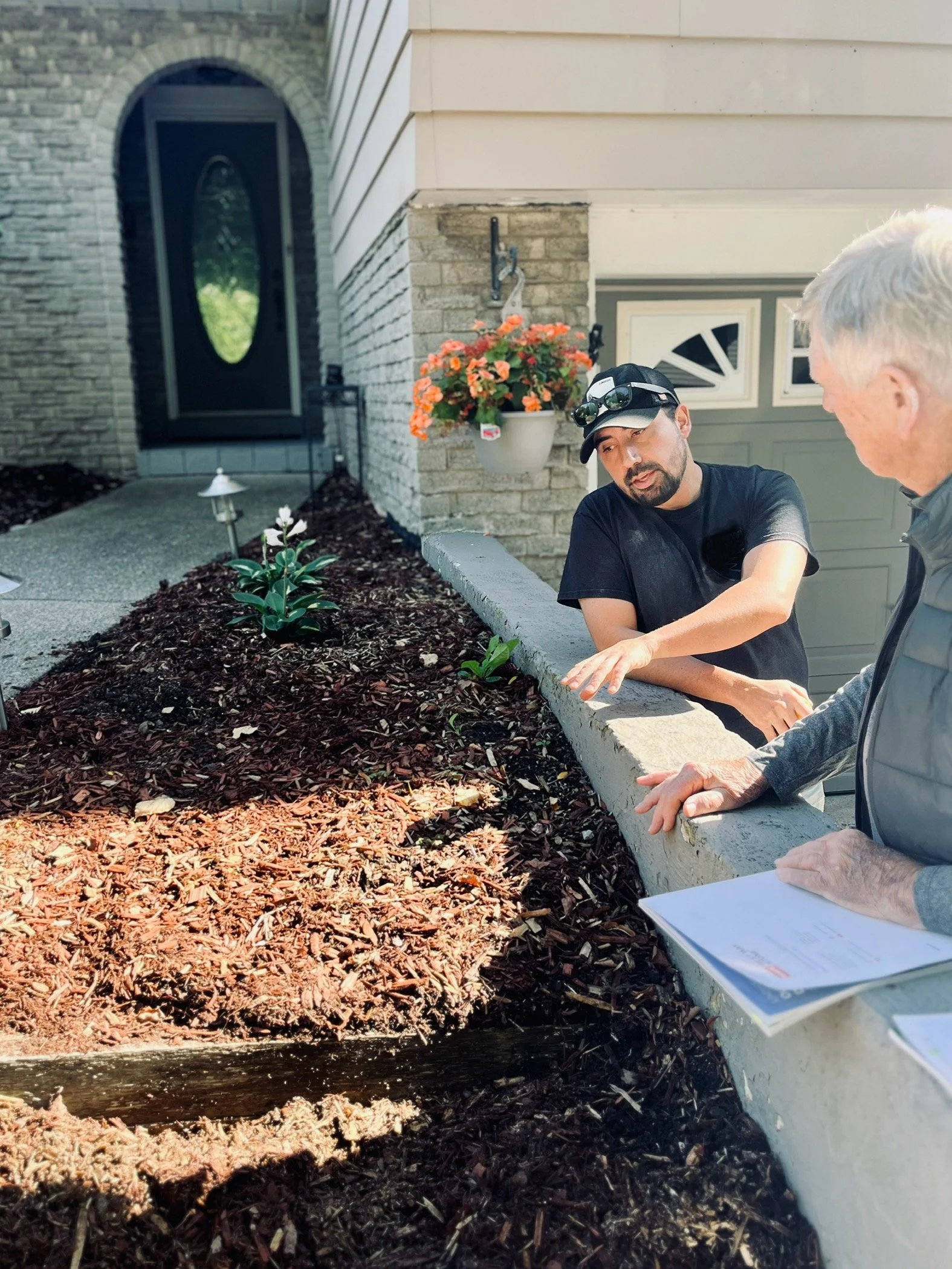 Two men are talking in front of a garden bed with flowers and mulch, near a house with a black front door and a garage. One man is holding papers and the other is gesturing.