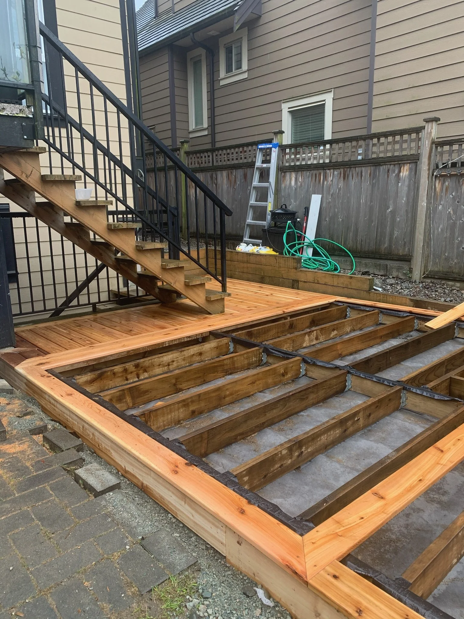 Image of a backyard deck construction project showing the wooden frame built on the ground, with future decking to be added, and a staircase leading up to the house. Tools and materials are visible in the background.