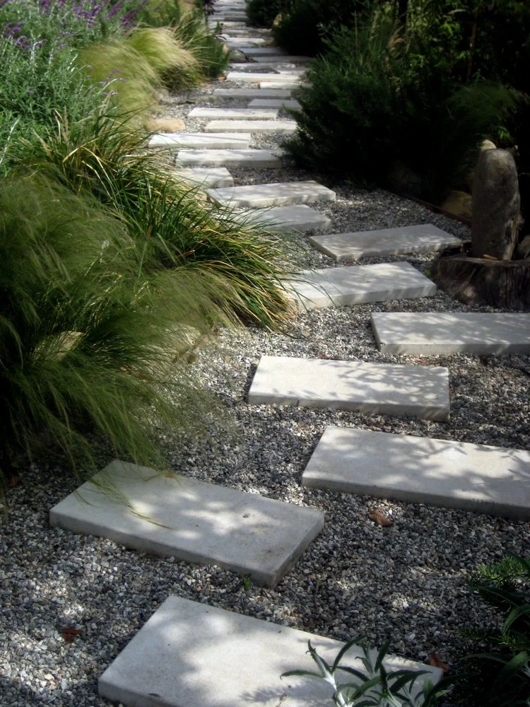 A garden pathway made of rectangular stepping stones surrounded by gravel, with green plants and bushes on each side.