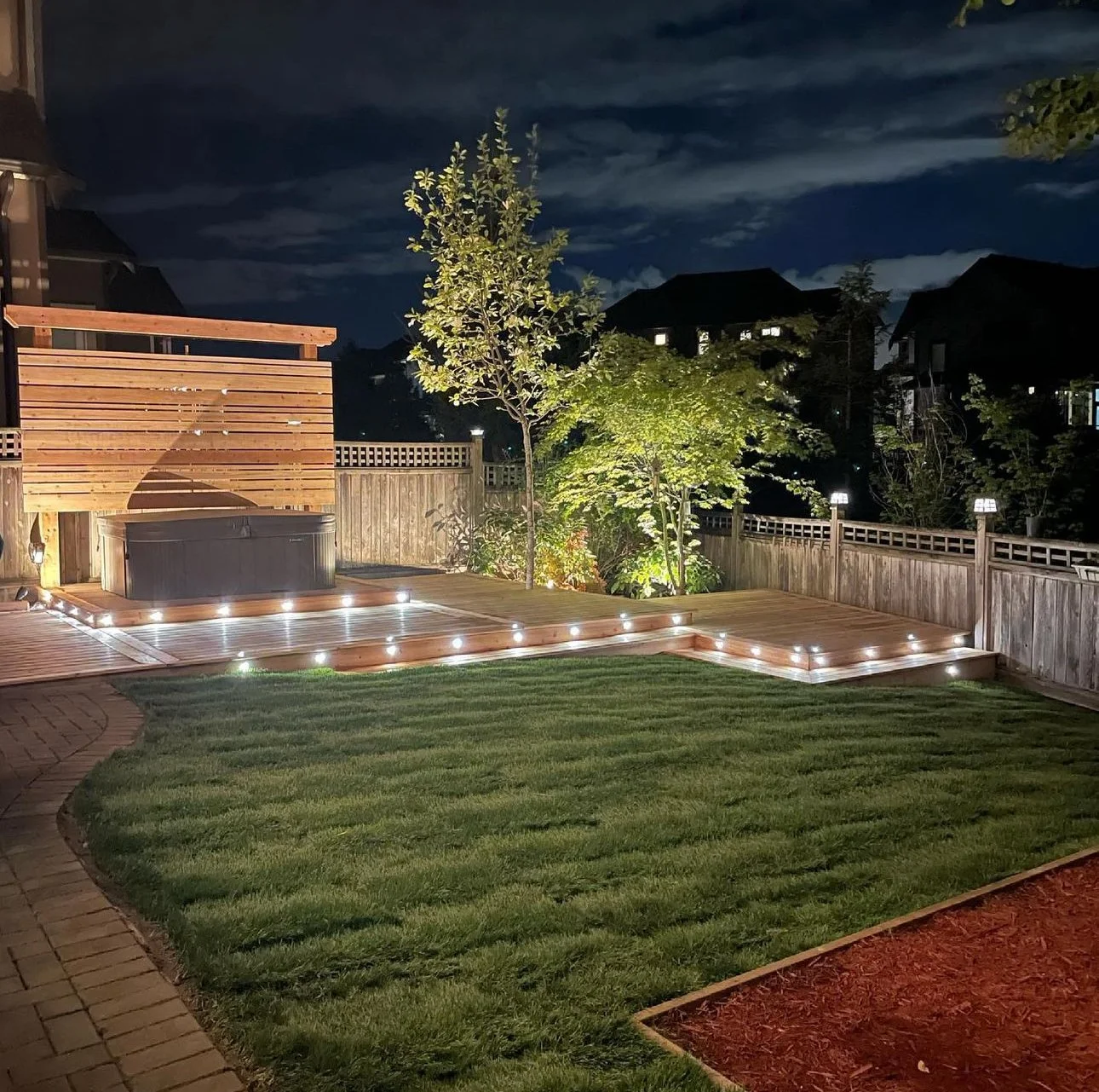 Nighttime backyard scene featuring a wooden deck with built-in LED lights surrounding a hot tub, a well-maintained lawn, a wooden fence, trees illuminated by outdoor lighting, and neighboring houses in the background.