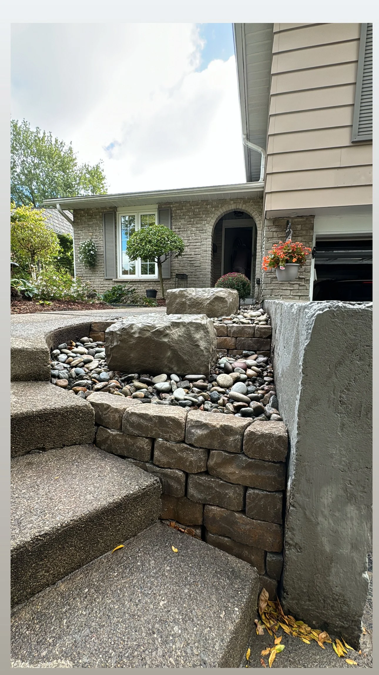Front yard with landscaped stone and brick retaining wall, large rocks, and a concrete staircase leading to the house entrance, with a well-maintained lawn and house with brick facade and window shutters.