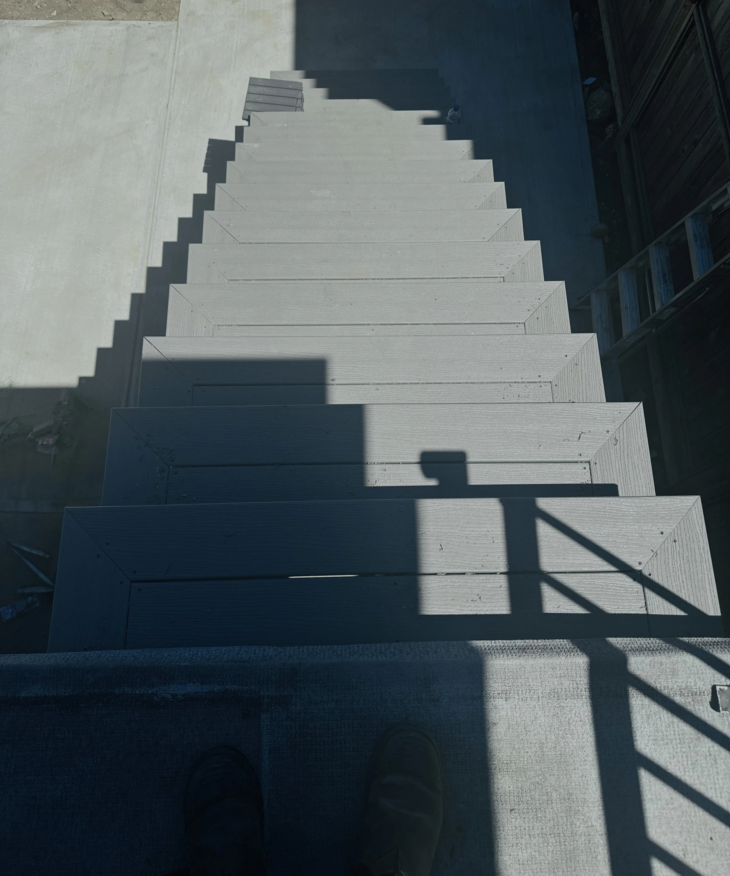 Looking down a set of outdoor stairs with a shadow cast by a railing and a street lamp on the steps, and a person's feet visible at the top of the stairs.