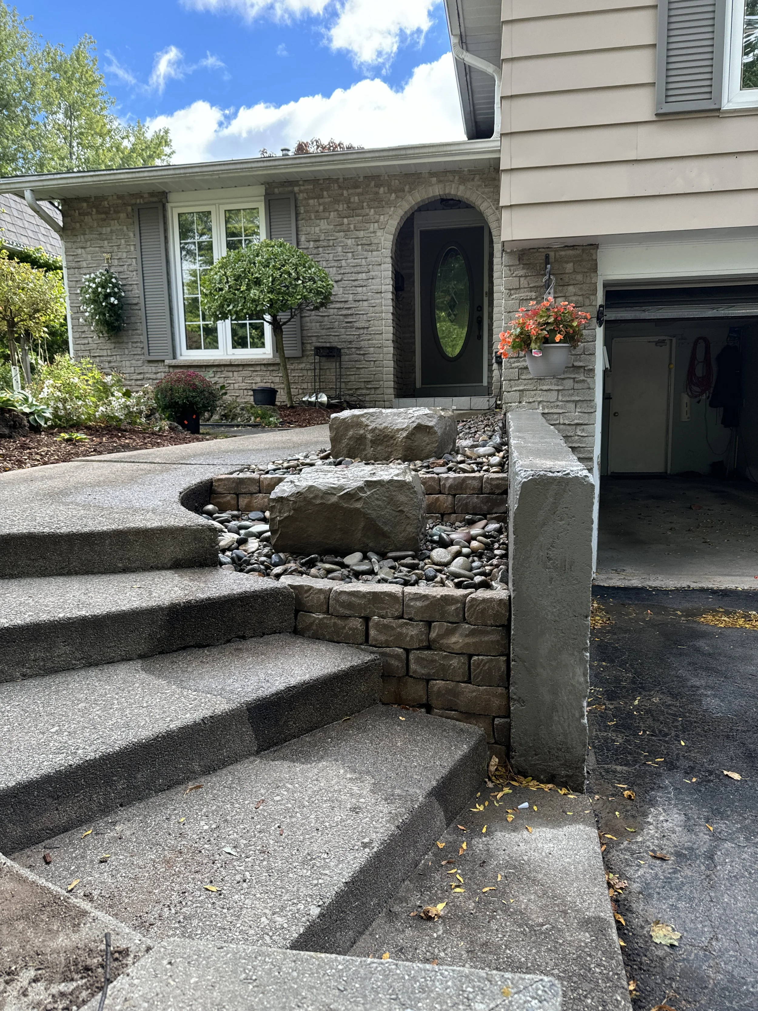 Front steps leading to a house with a landscaped yard, brick walls, and a circular window on the front door. A flower pot with pink flowers hangs beside the garage door.