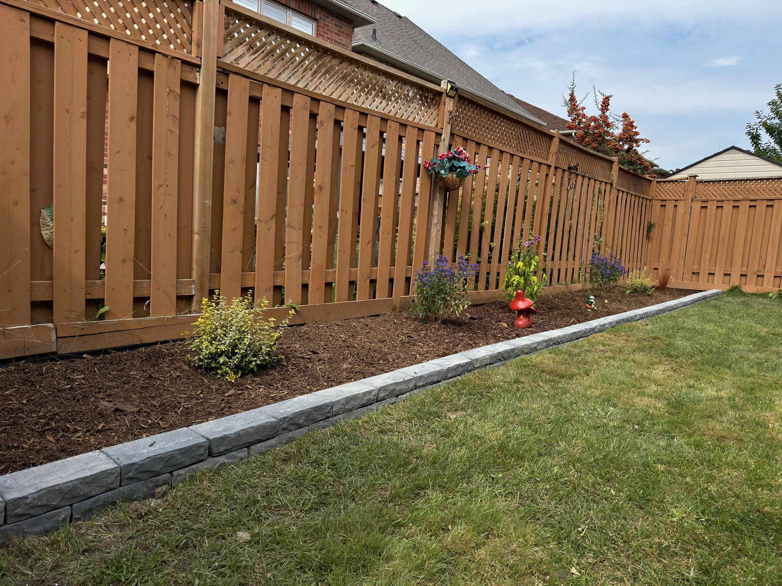 A backyard garden with a wooden fence, flower bed with small plants, decorative garden ornaments, and a stone edging, with grass in the foreground.