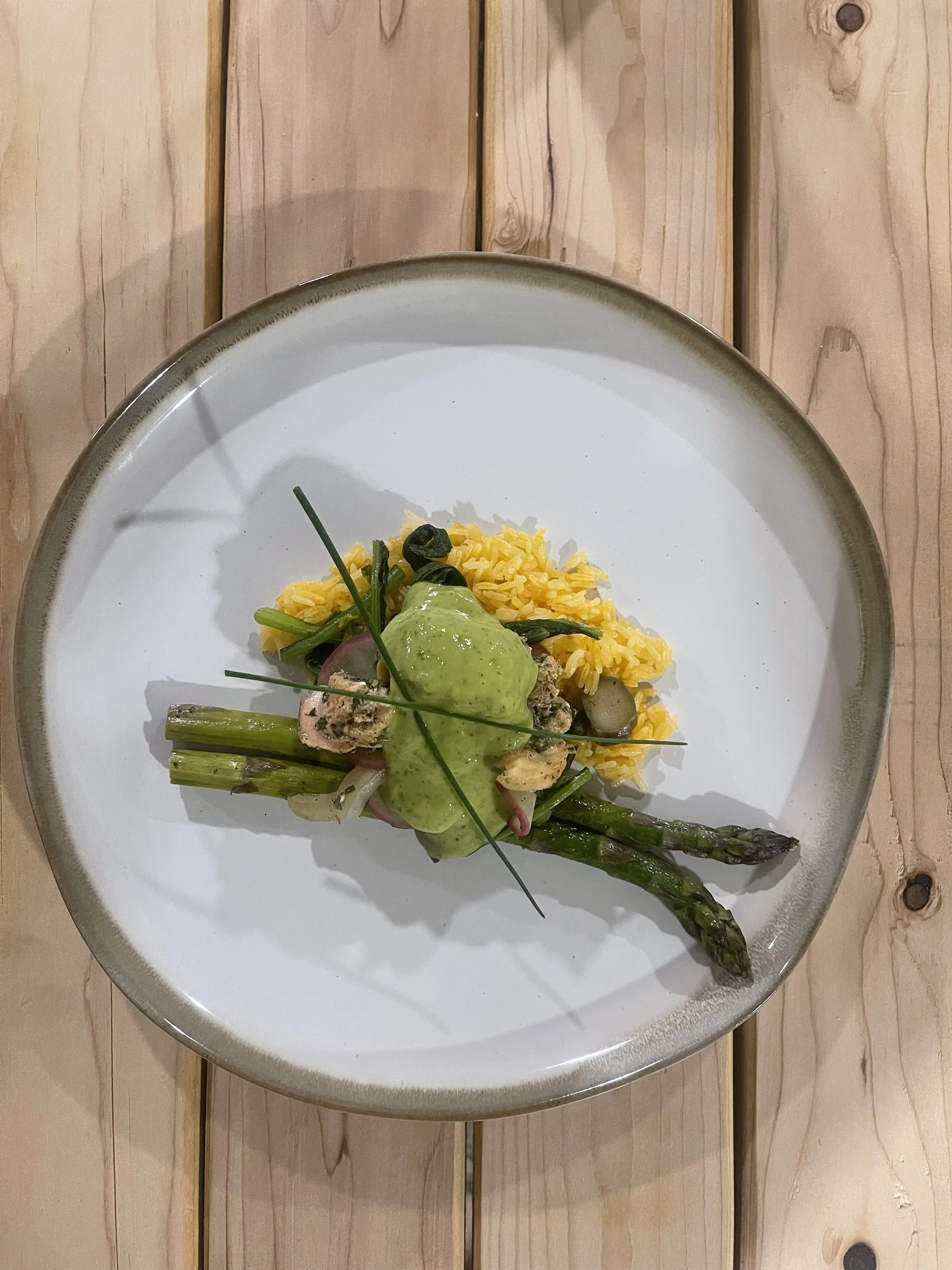 A white plate with rice and grilled vegetables, topped with green sauce, placed on a wood table.