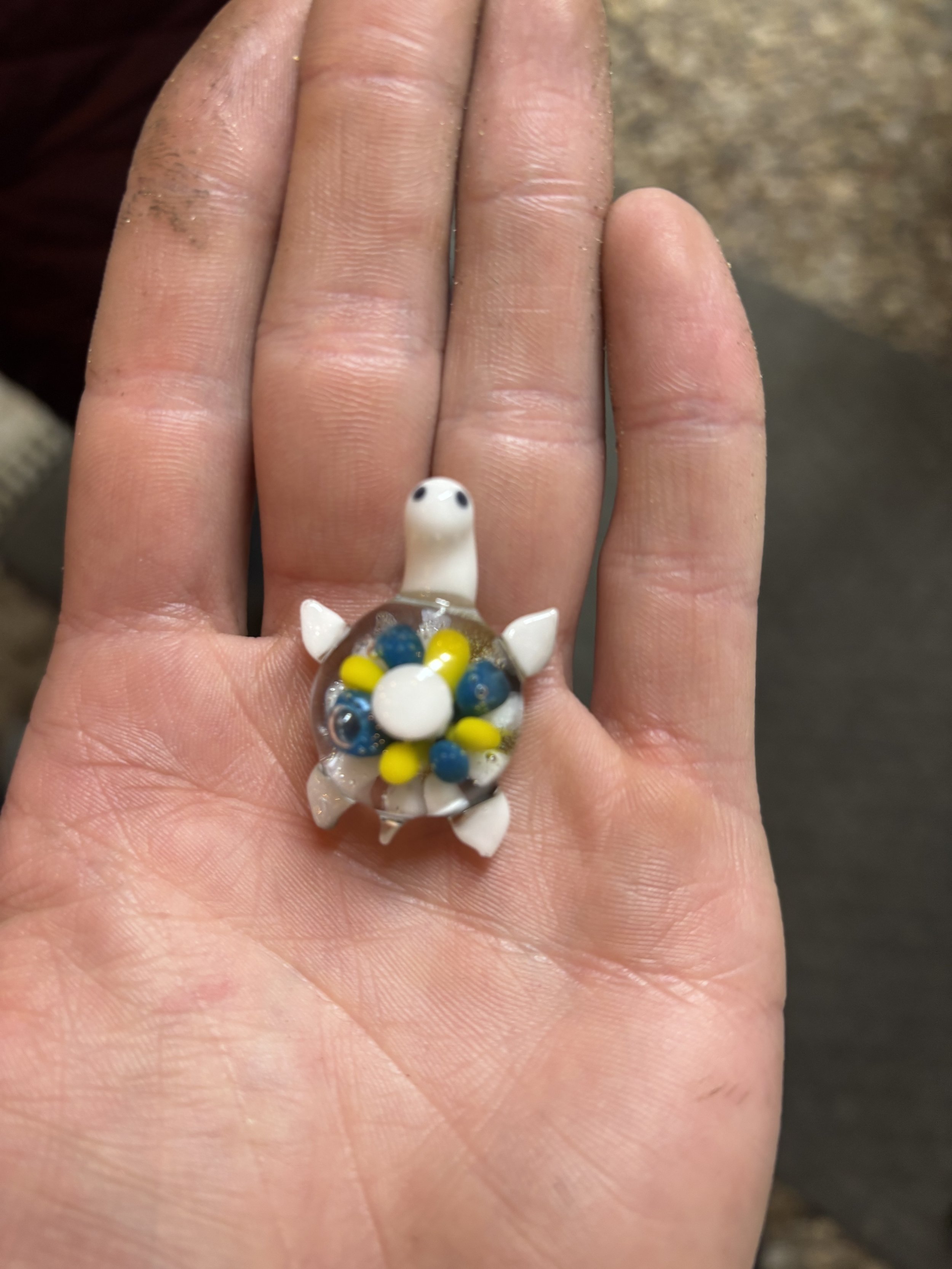 A small glass turtle figurine with a white head and limbs, and a shell filled with blue, yellow, and clear beads, resting on a person's open palm.
