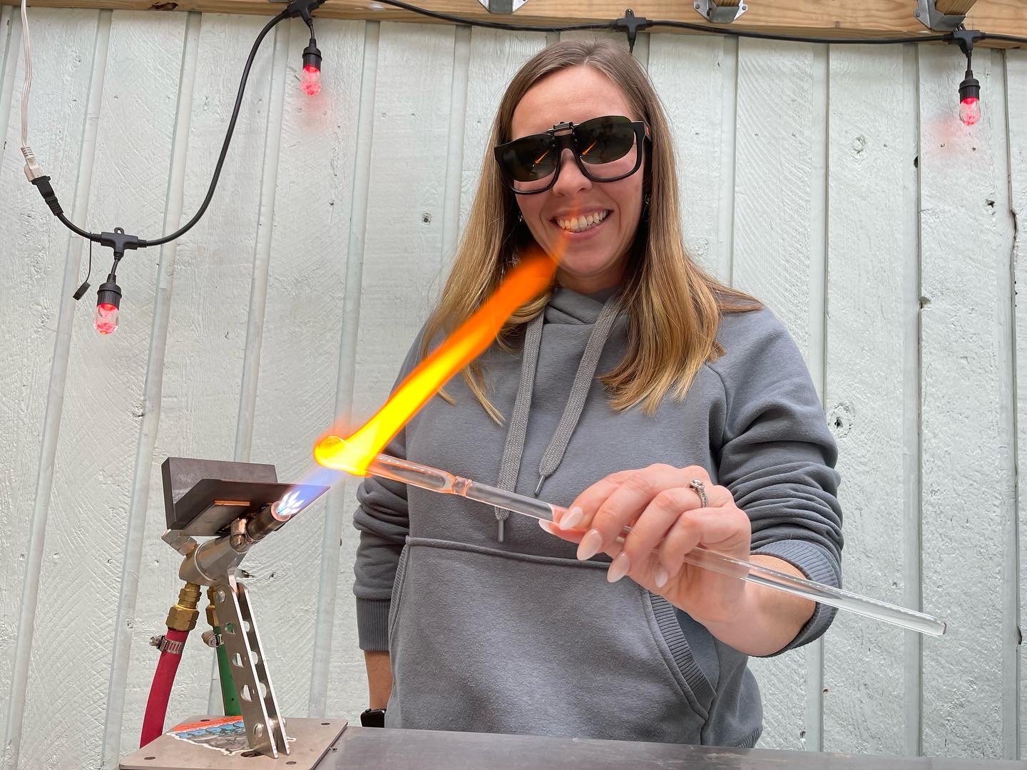 Woman with sunglasses smiling while holding a glass tube with orange flames, working at a glassblowing setup outdoors.