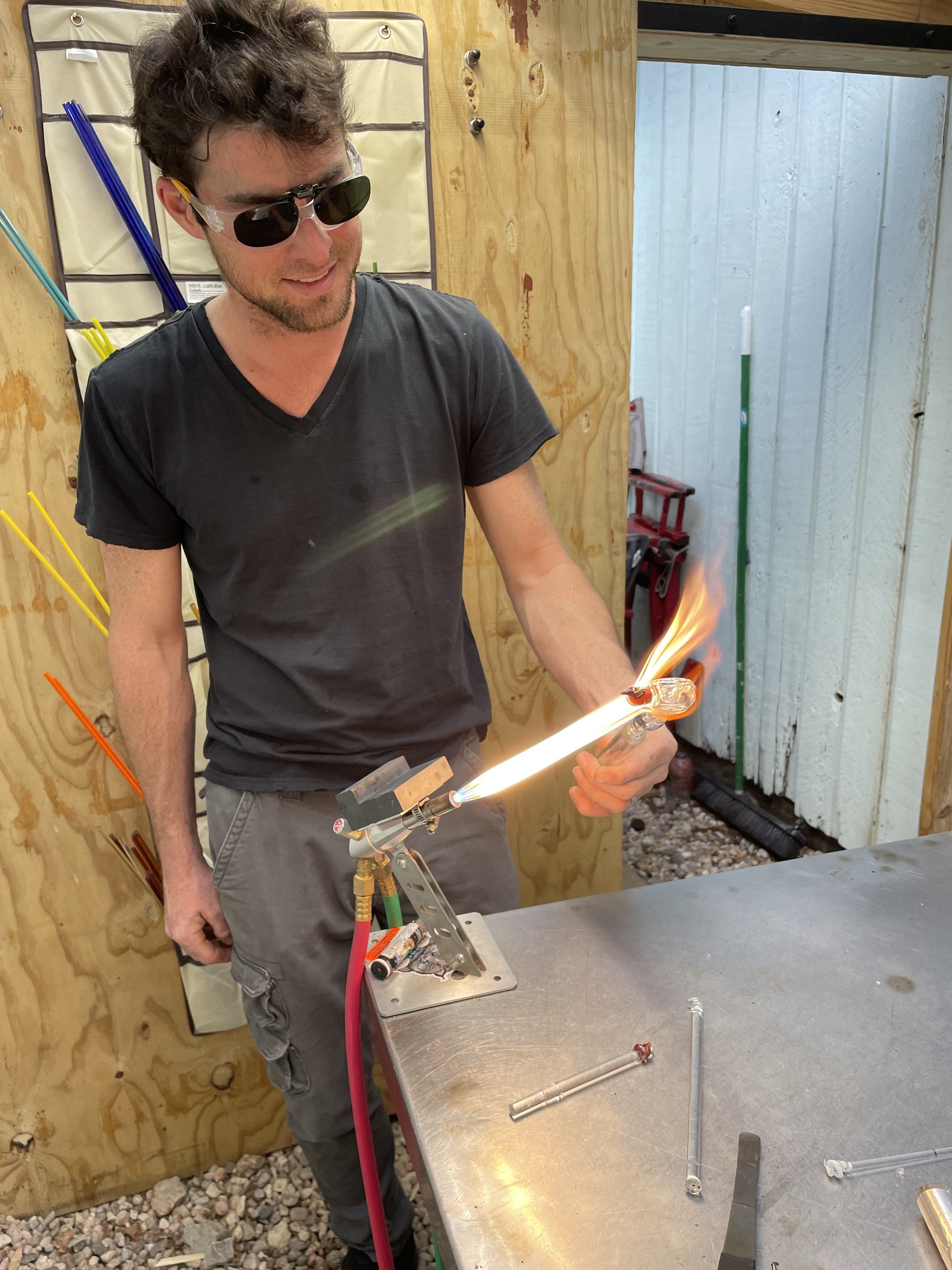 A man wearing sunglasses and a black t-shirt is using a blowtorch to work on glass or a similar material in a workshop. He is smiling and focused on his task.