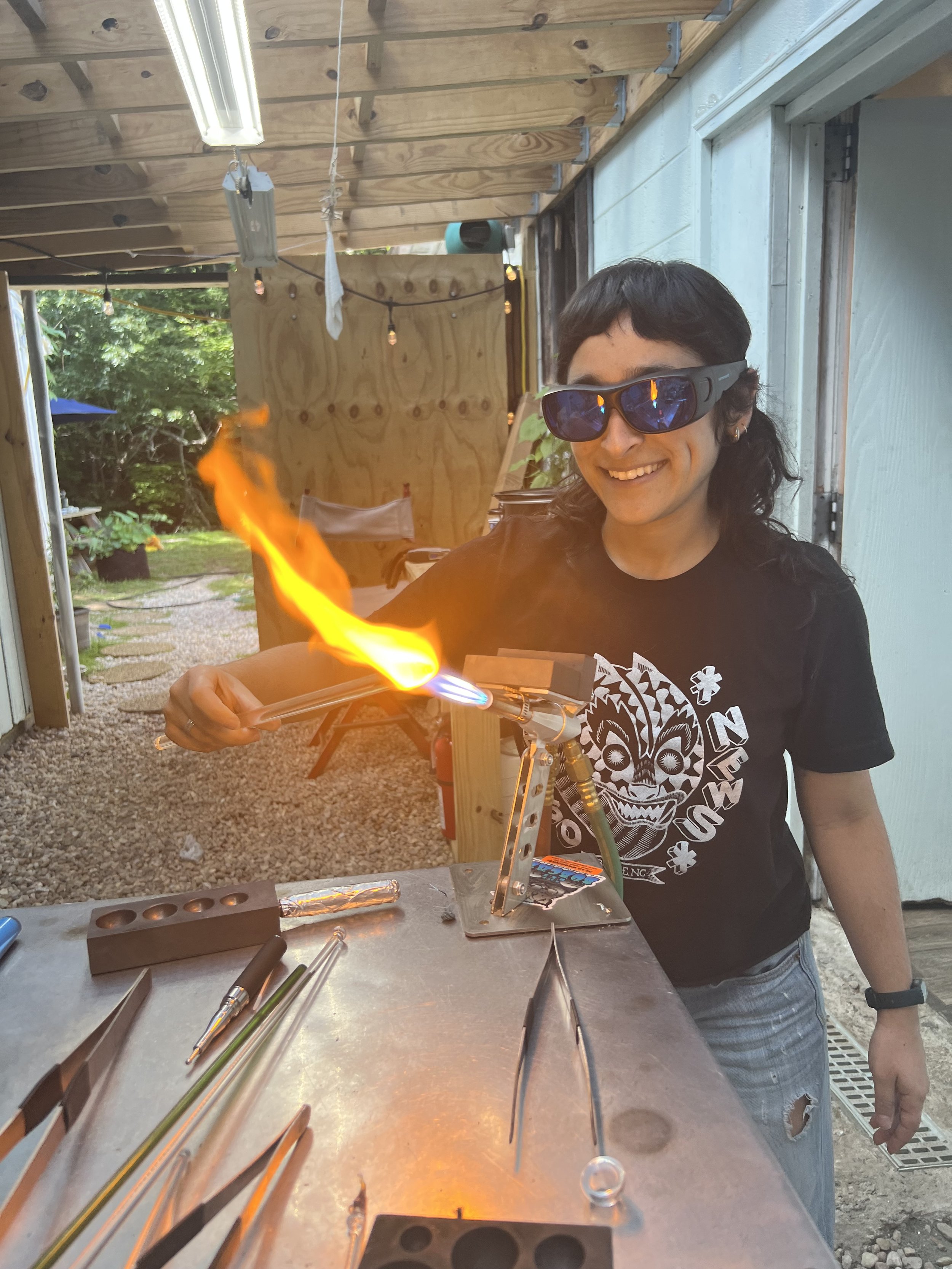 Person wearing sunglasses and a black T-shirt, smiling, working with a flame torch on a glassworking or metalworking project in a workshop outdoors.