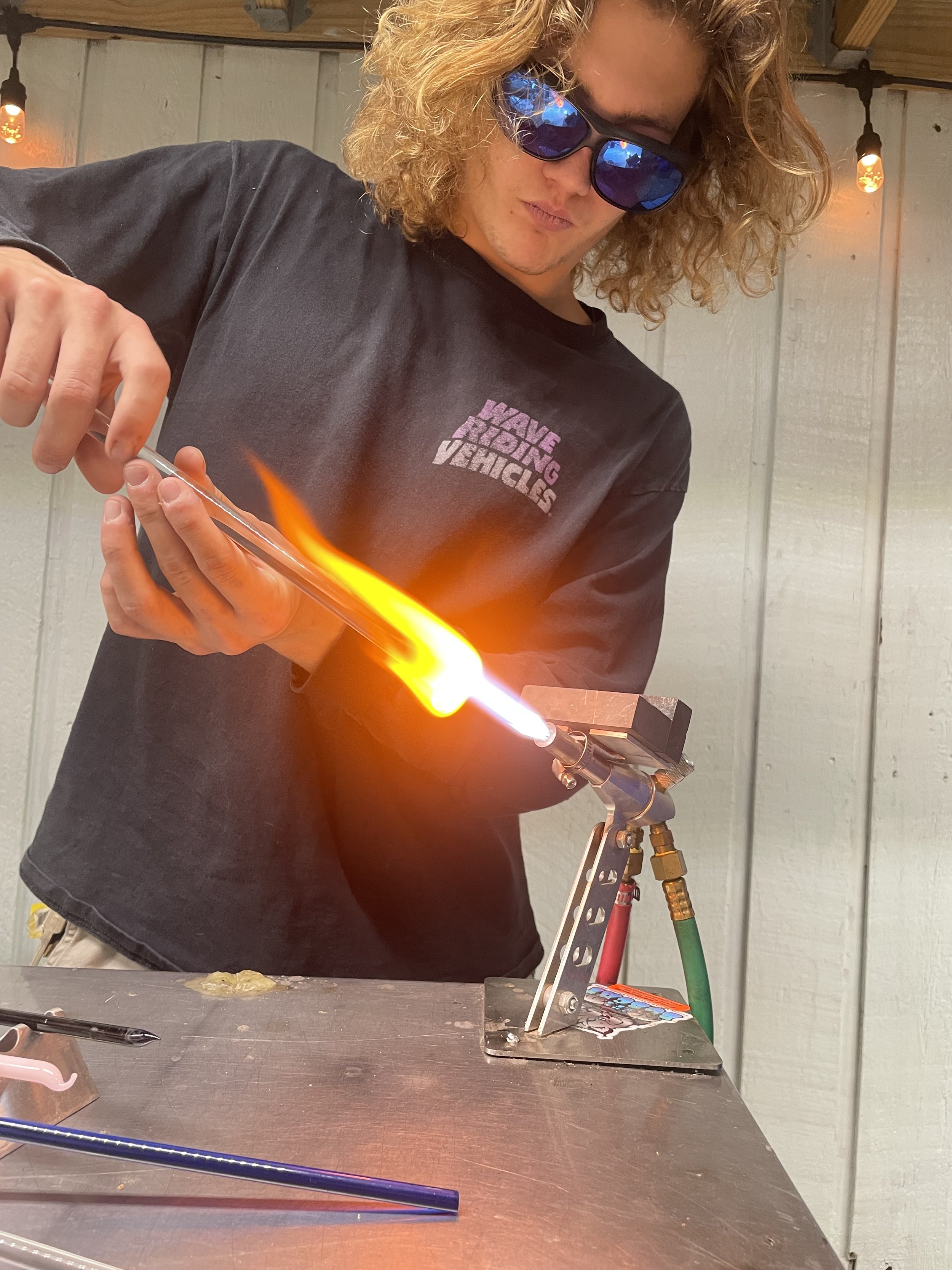 A person with curly blonde hair wearing sunglasses and a black shirt that says 'Have a Nice Day' is performing a glassblowing process, shaping molten glass with a torch in a workshop setting.
