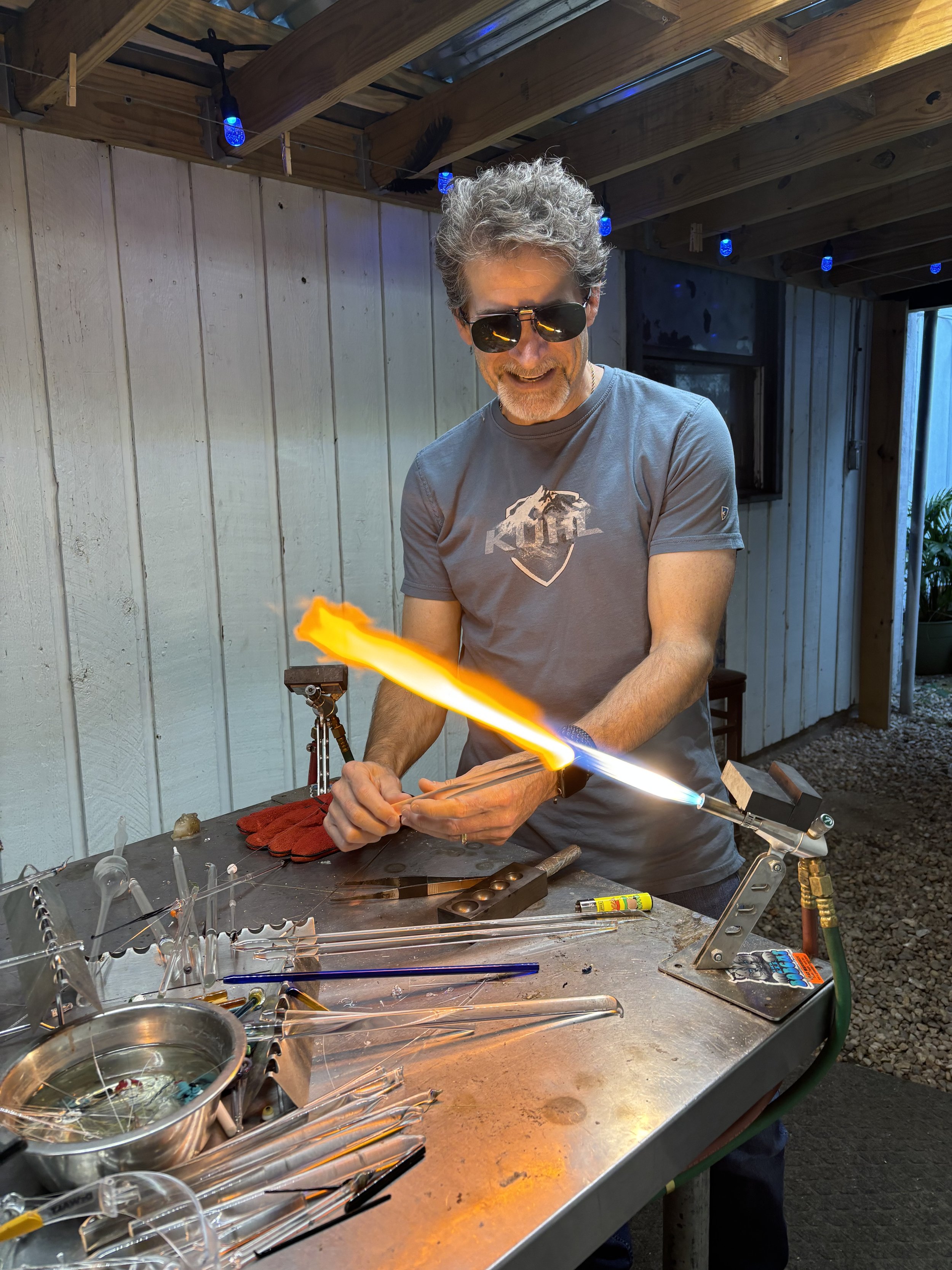 A man with sunglasses and a gray T-shirt is working in a glassblowing studio, using a torch to shape glass. The worktable in front of him holds various glassblowing tools, glass tubes, and a bowl of water. The background includes a wooden wall and ce