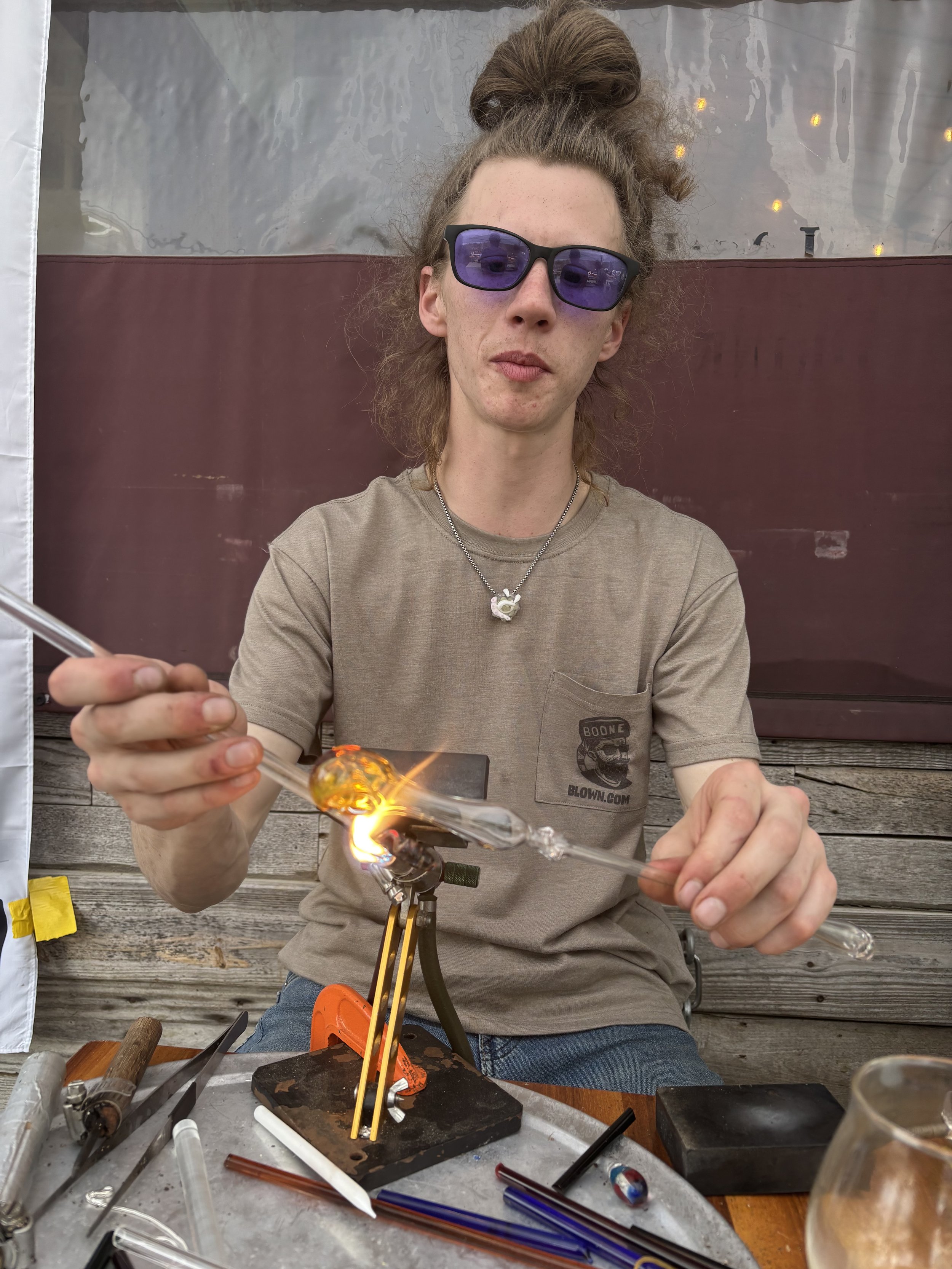 A man with curly hair in a bun, wearing sunglasses and a beige t-shirt, is working with a glassblowing tool over a small torch flame, creating glass art. She is seated at a workbench with various glassblowing tools and materials around her.