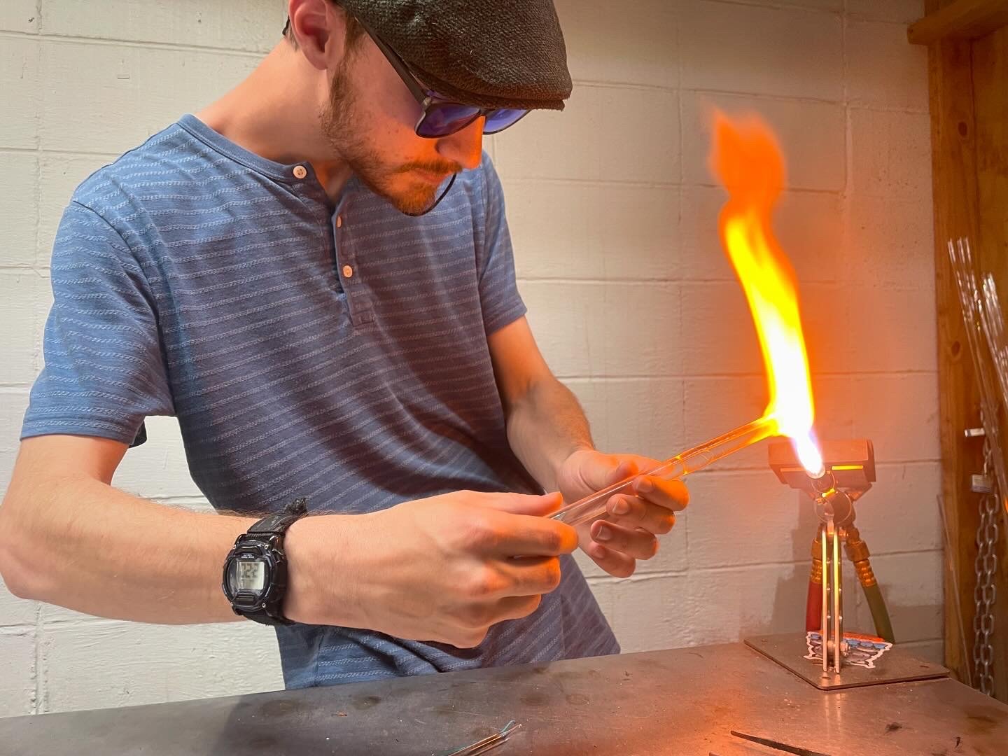 Man in a blue shirt and glasses working with glassblowing, heating glass with a torch that produces a tall flame.