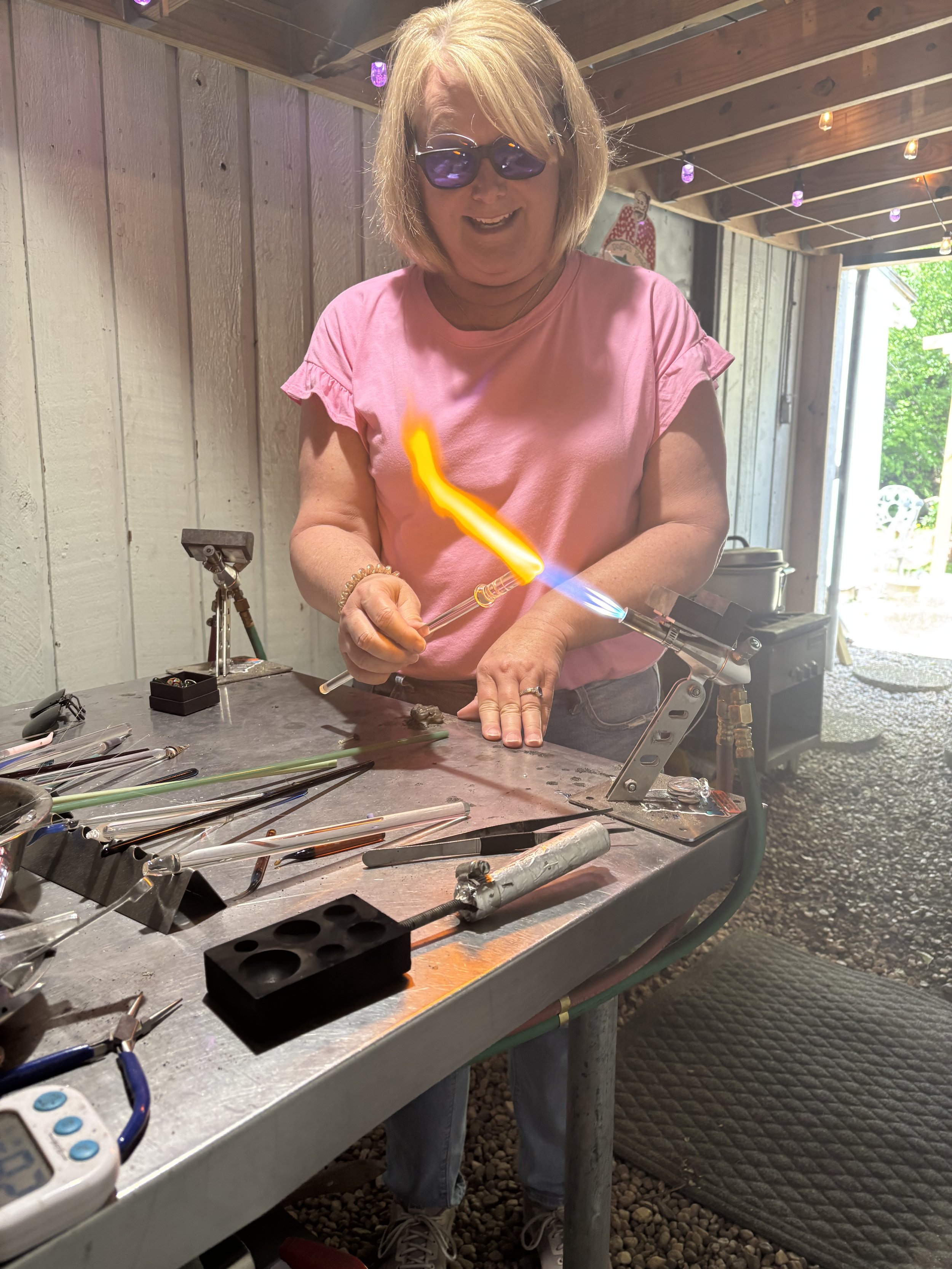 A woman wearing sunglasses and a pink shirt is working on glassblowing, making a glass object with a torch in a workshop.