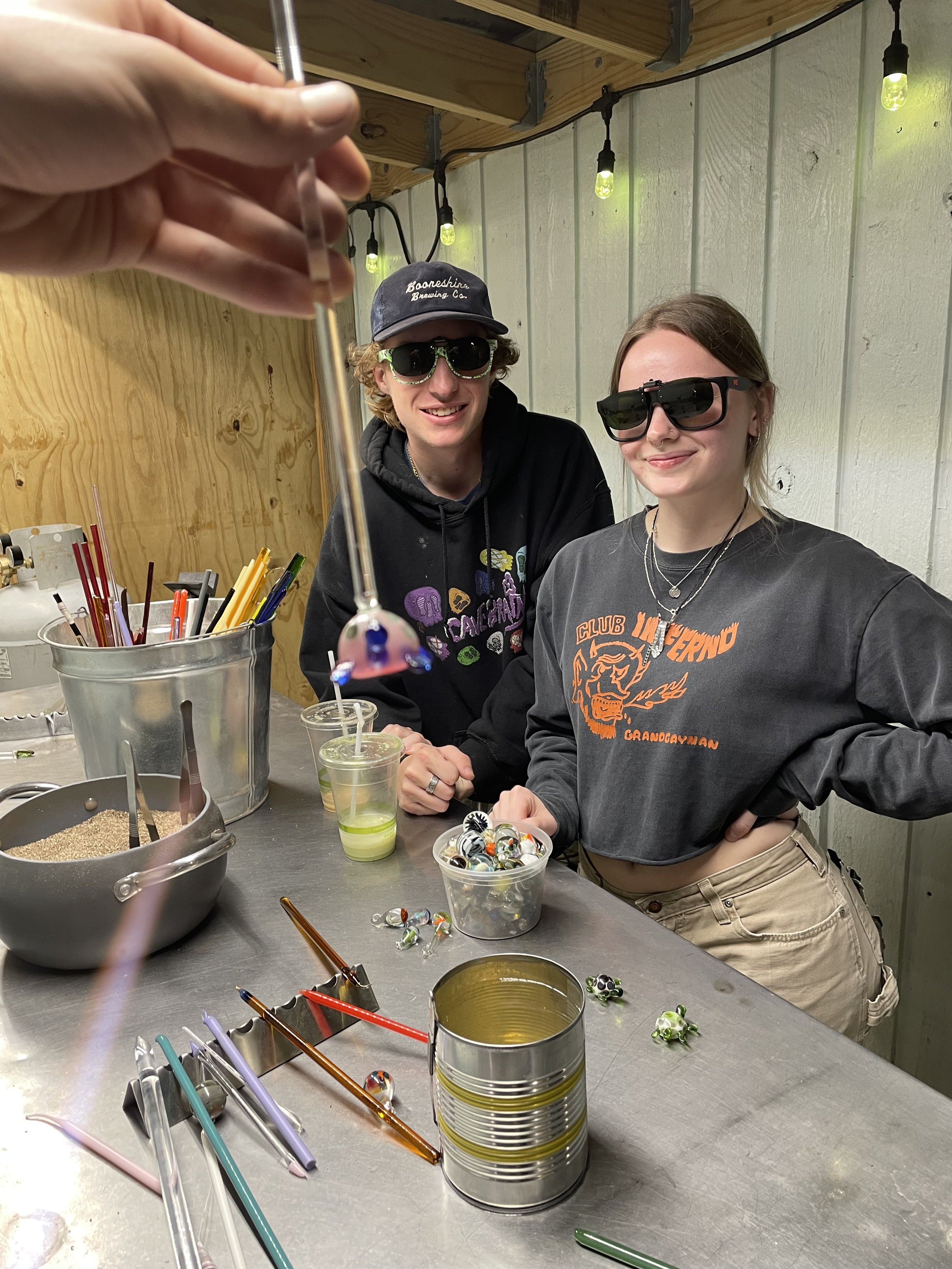 Two people wearing sunglasses smiling at a craft table, with a metal can, colorful glass marbles, and craft supplies scattered on the table.