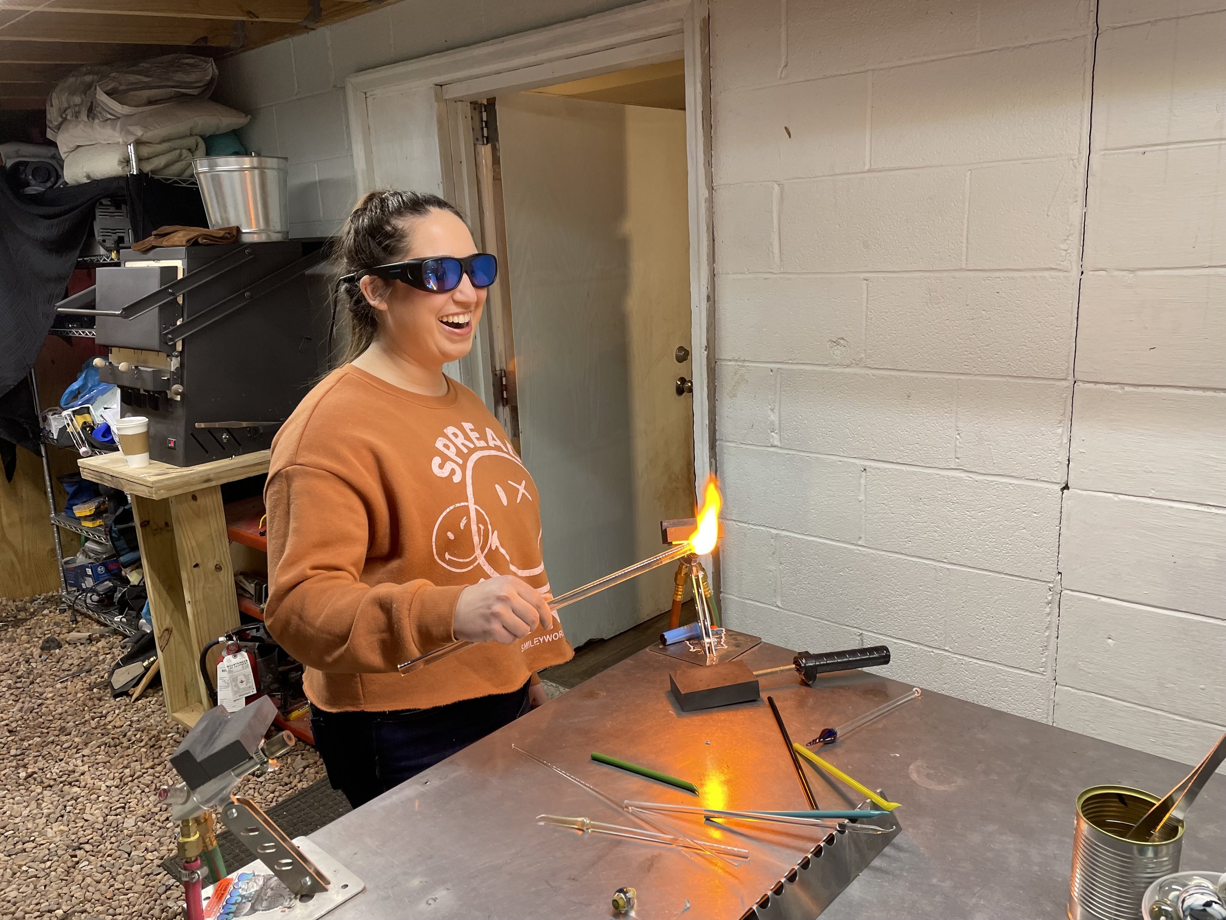 A woman wearing safety glasses smiles while working with a flame on a glass or metal rod in a workshop, surrounded by tools and glass rods on a metal table.