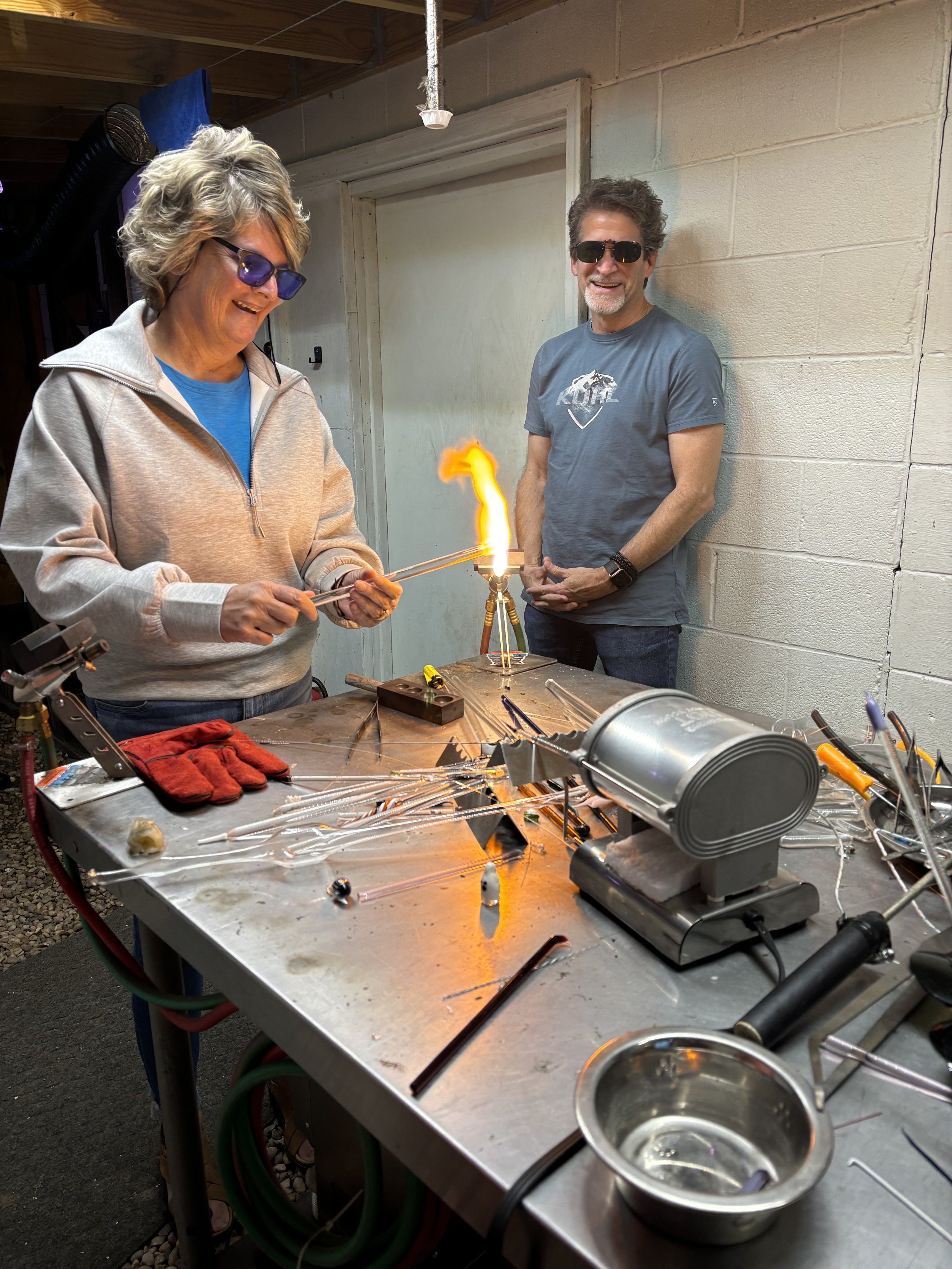 Two people, a woman and a man, are glassblowing together in a workshop. The woman is holding a glowing glass piece with tongs, and the man is smiling while observing. The worktable has various glassblowing tools and equipment, with a flame in the cen