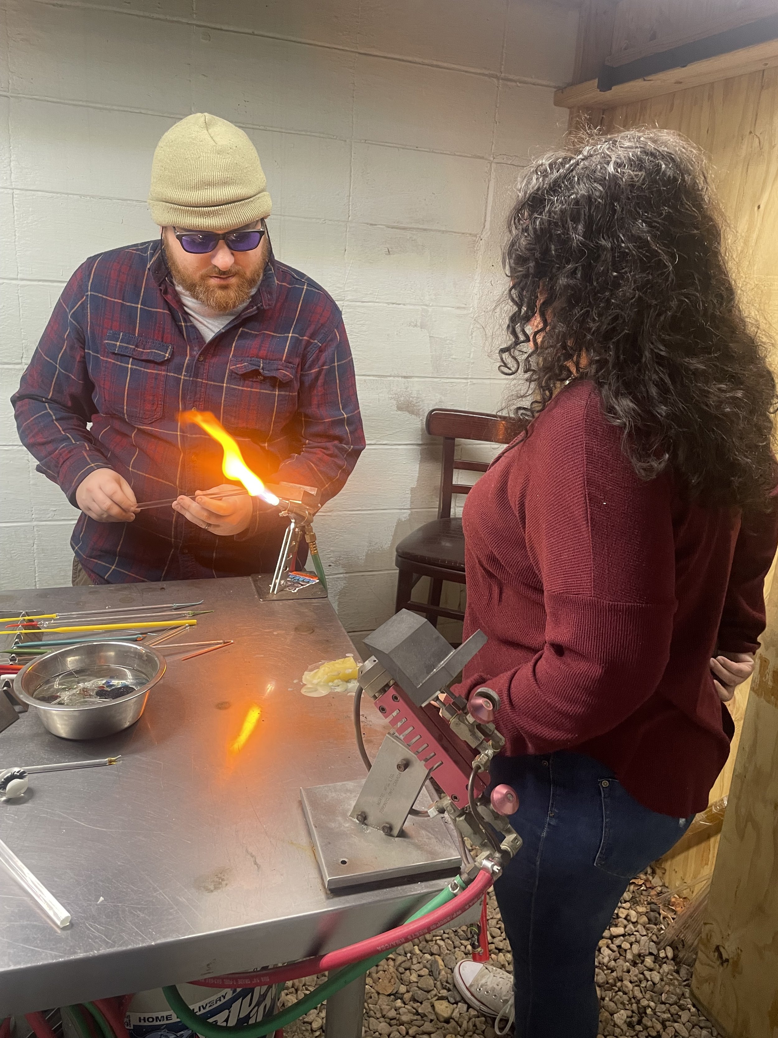A man and woman practicing glass blowing. The man is wearing a beige beanie, sunglasses, and a red plaid shirt, holding a glass piece with a flame nearby. The woman is wearing a maroon sweater and jeans, observing. Glassworking tools and supplies are