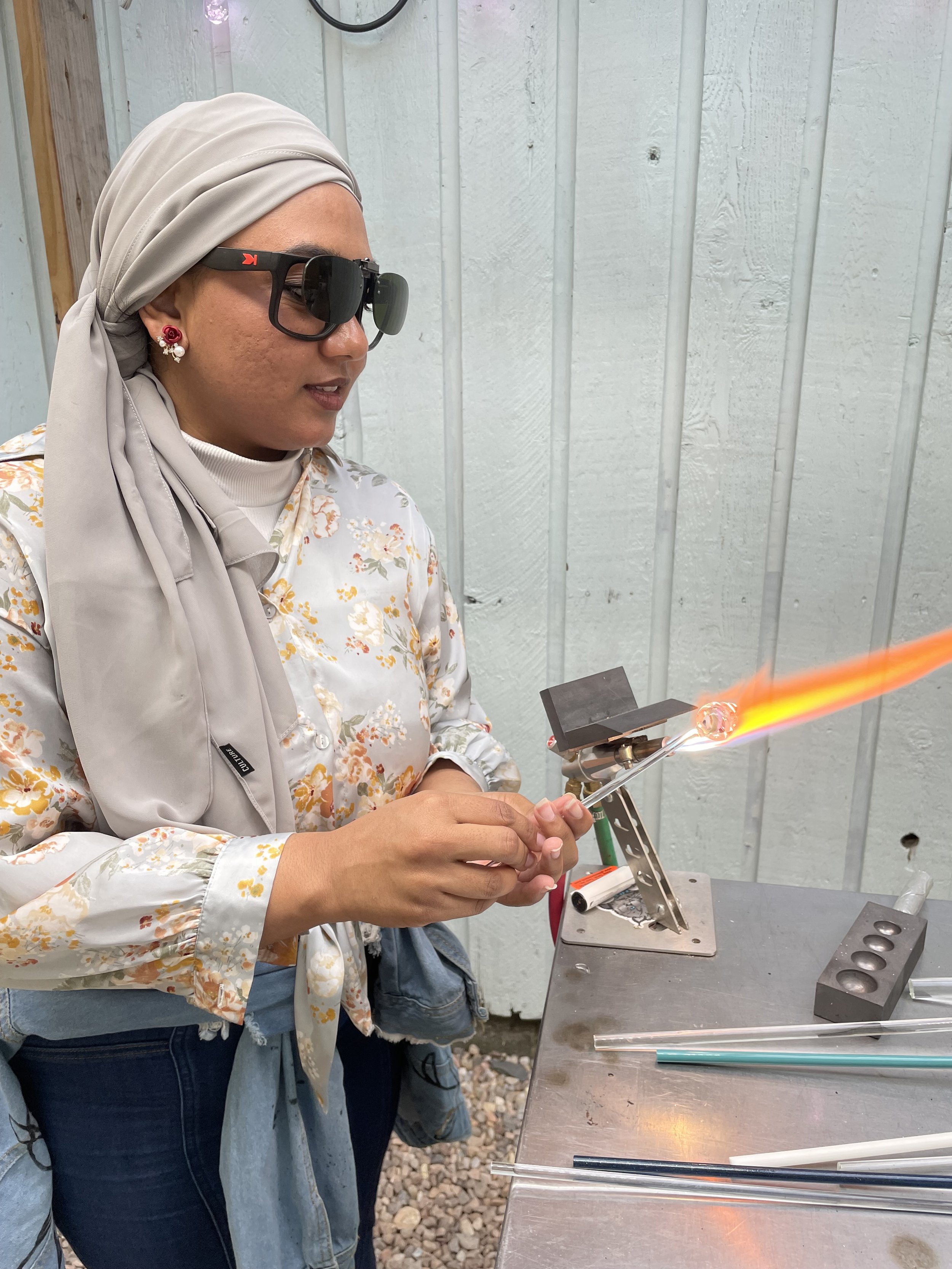 A woman wearing a headscarf, sunglasses, and floral blouse is working with glassblowing tools, shaping a glass piece with flame in a workshop setting.
