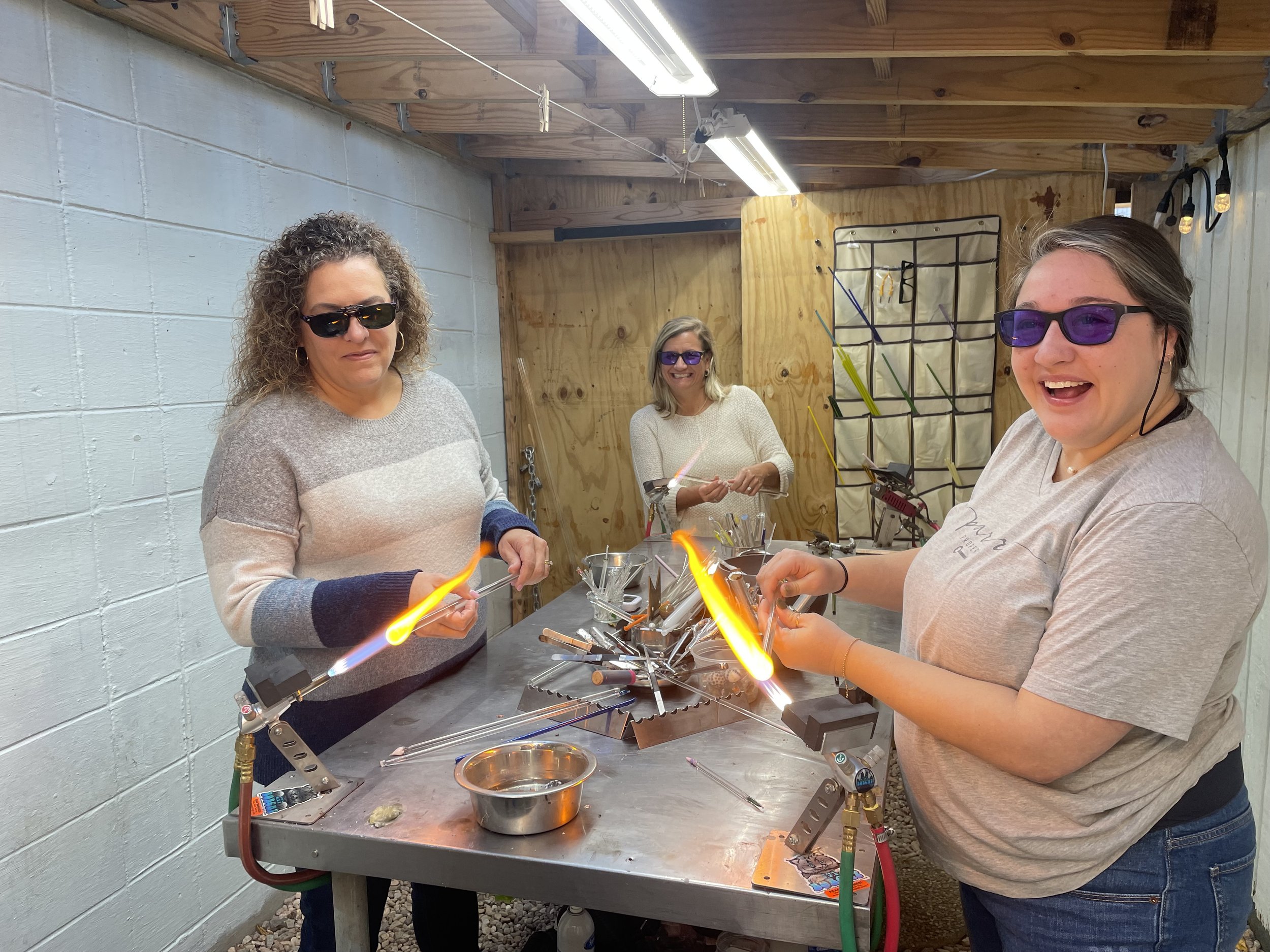 Three women wearing safety glasses are glassblowing at a workbench with tools and glowing glass rods in a workshop.
