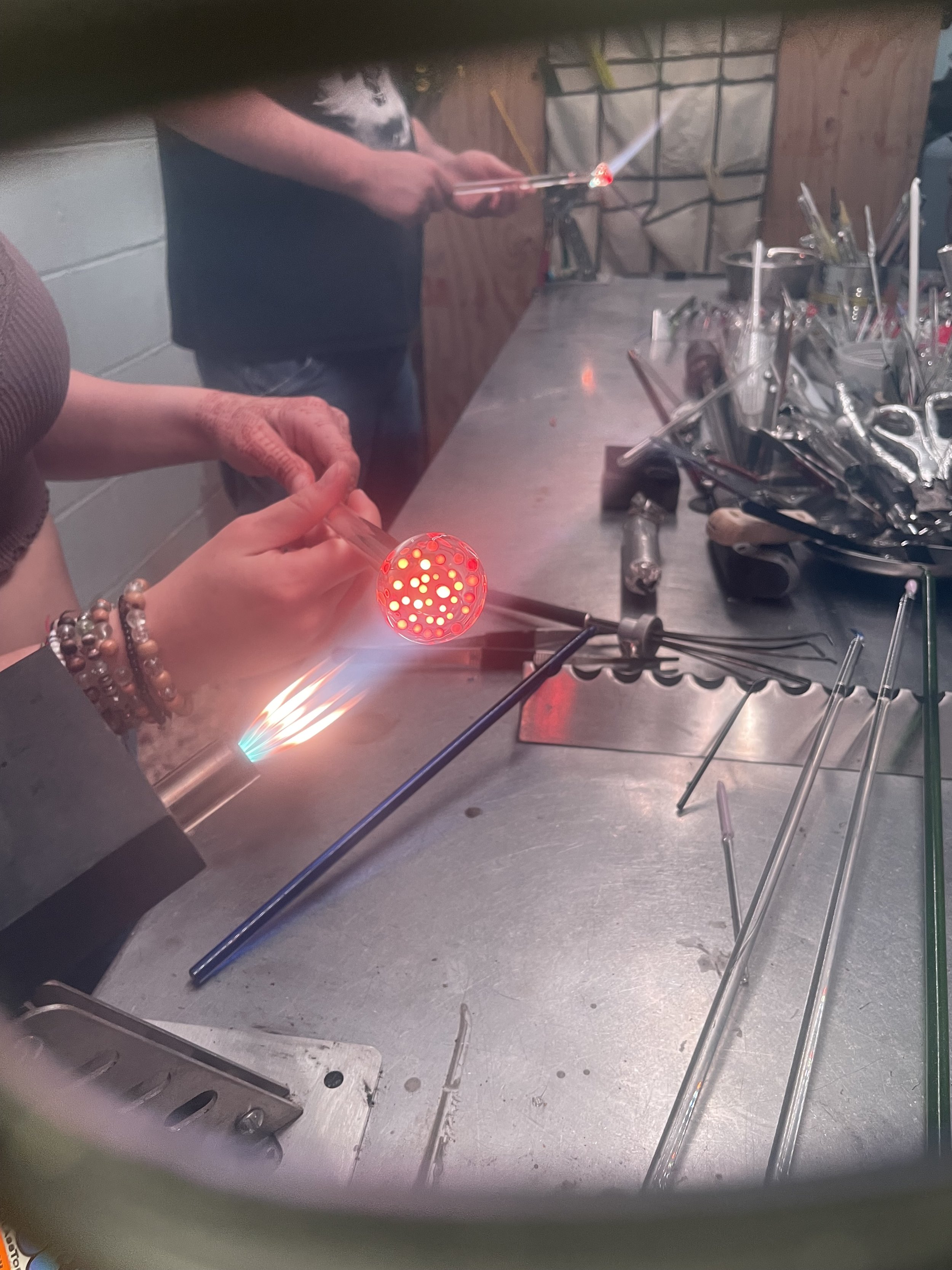 A person is working on glass blowing, holding a heated glass piece with tongs in a workshop. The glass is glowing red and has holes, with a small torch directed at it. Various glassblowing tools are on the work surface nearby.