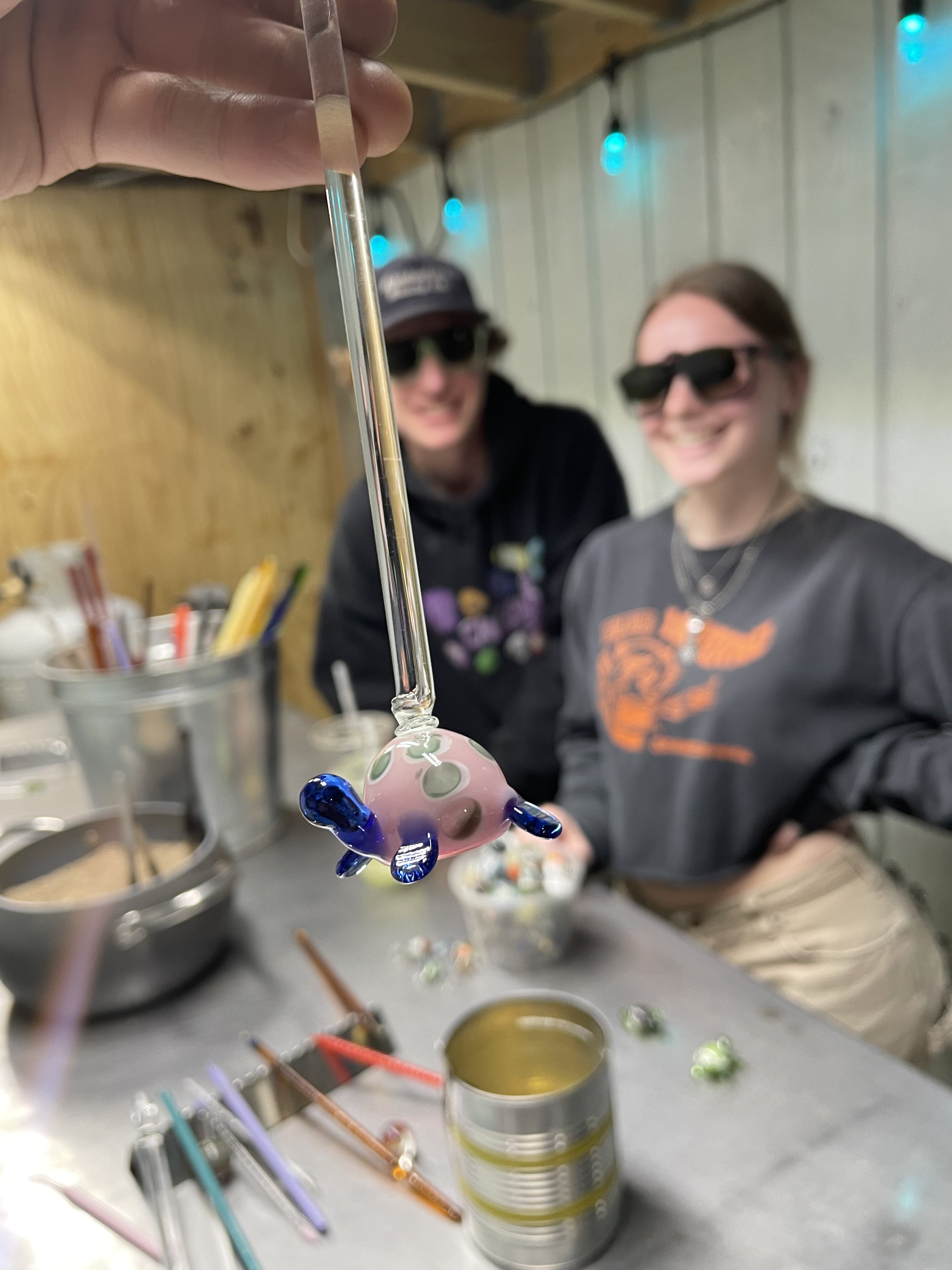 A glass pacifier-shaped object with blue and pink colors being held up by a person. Two women in sunglasses and casual clothing are smiling in the background indoors with string lights and wooden panel walls.